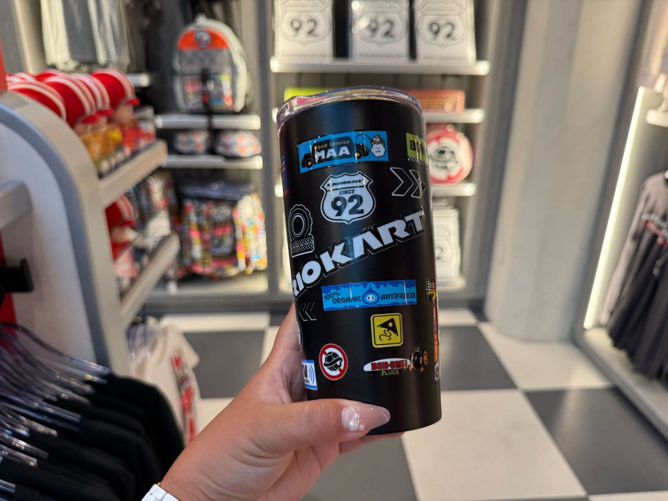 A hand holds a black Mario Motors tumbler in a Super Nintendo World shop, shelves behind—no castle like Disney's iconic park designs.