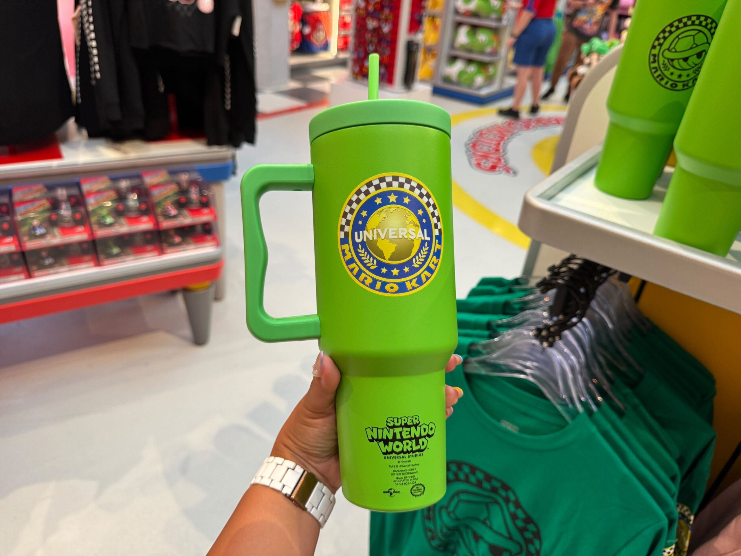 A hand holds a large green Mario Kart cup in a gift shop with Super Nintendo World items, evoking Disney parks’ immersive merch shops.