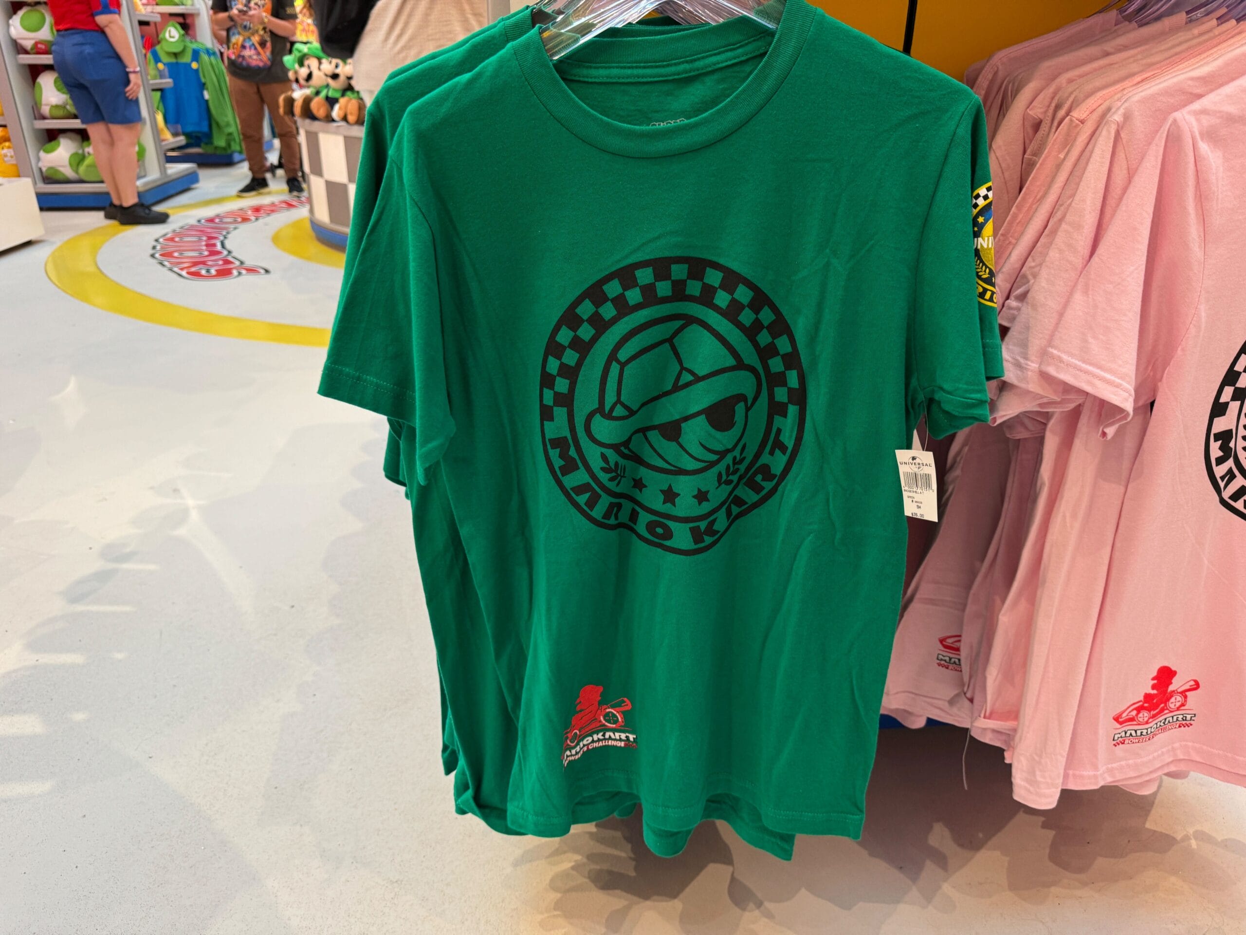 A green Mario Kart t-shirt featuring a black racing helmet, like theme park merch at Disney World, hangs on a store rack.