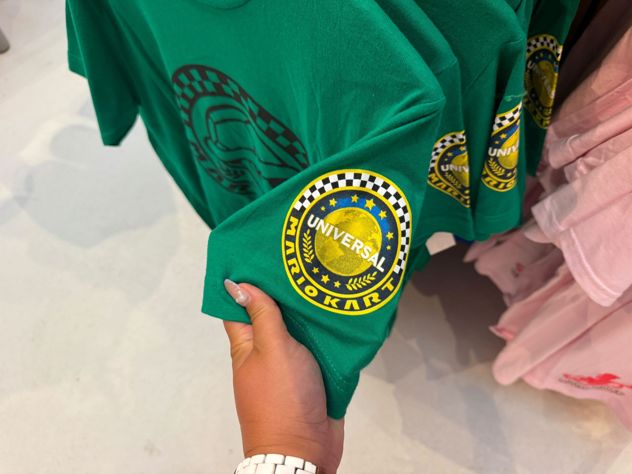 A hand holds a green "Universal Mario Kart" shirt—unlike Disney Parks castle tees, this features Mario Motors merchandise.