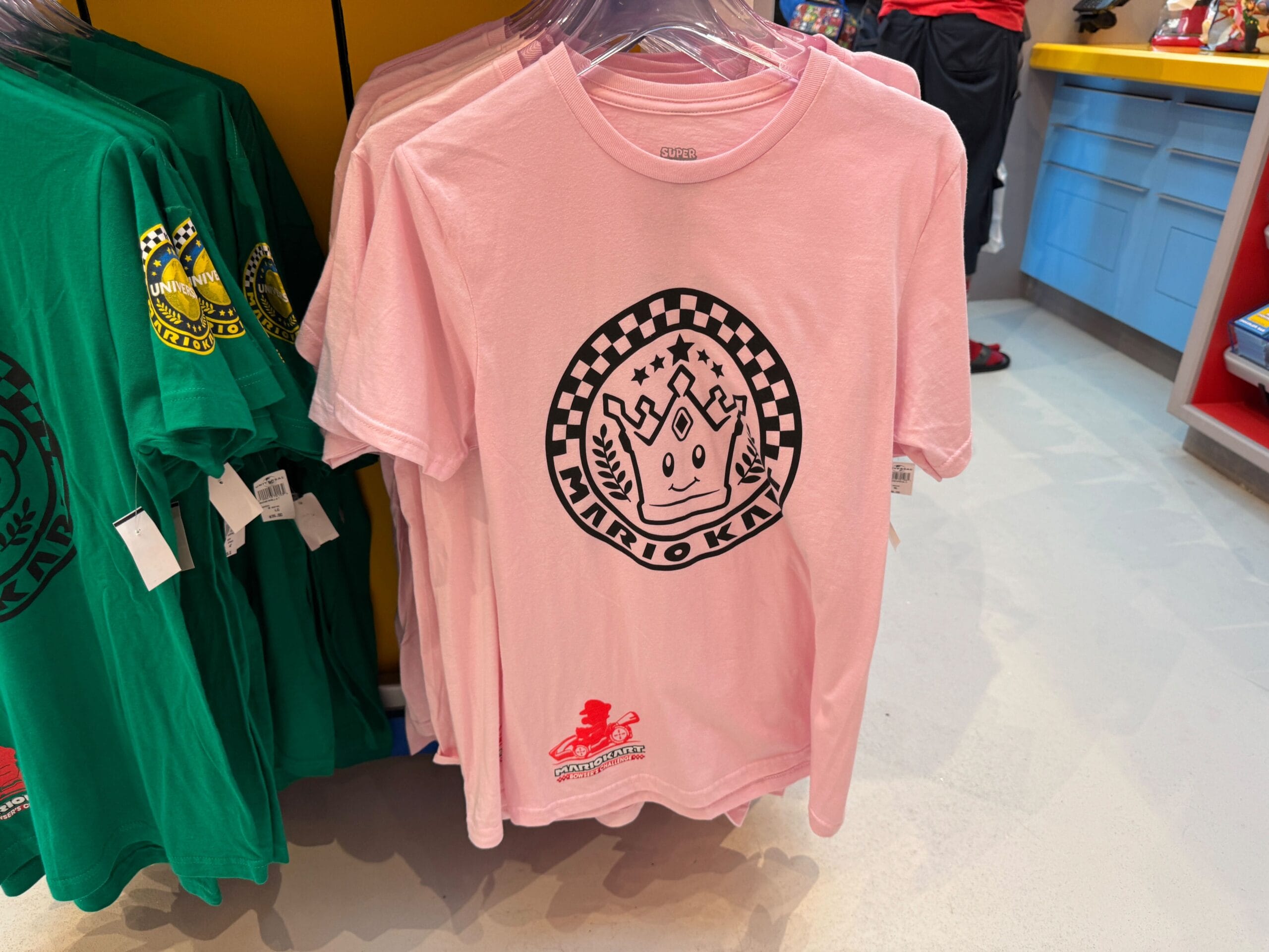 A pink Mario Kart T-shirt with a crowned logo, part of Mario Motors merch, is on a rack—no Disney castle like at the parks.