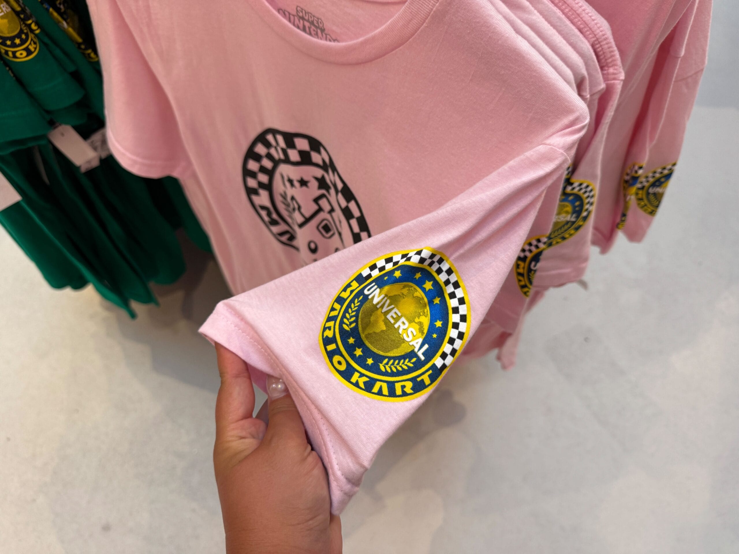 Hand holding a pink "Universal Kart to Kart" tee, with Mario Motors merchandise stacked nearby—not a Disney Parks castle.