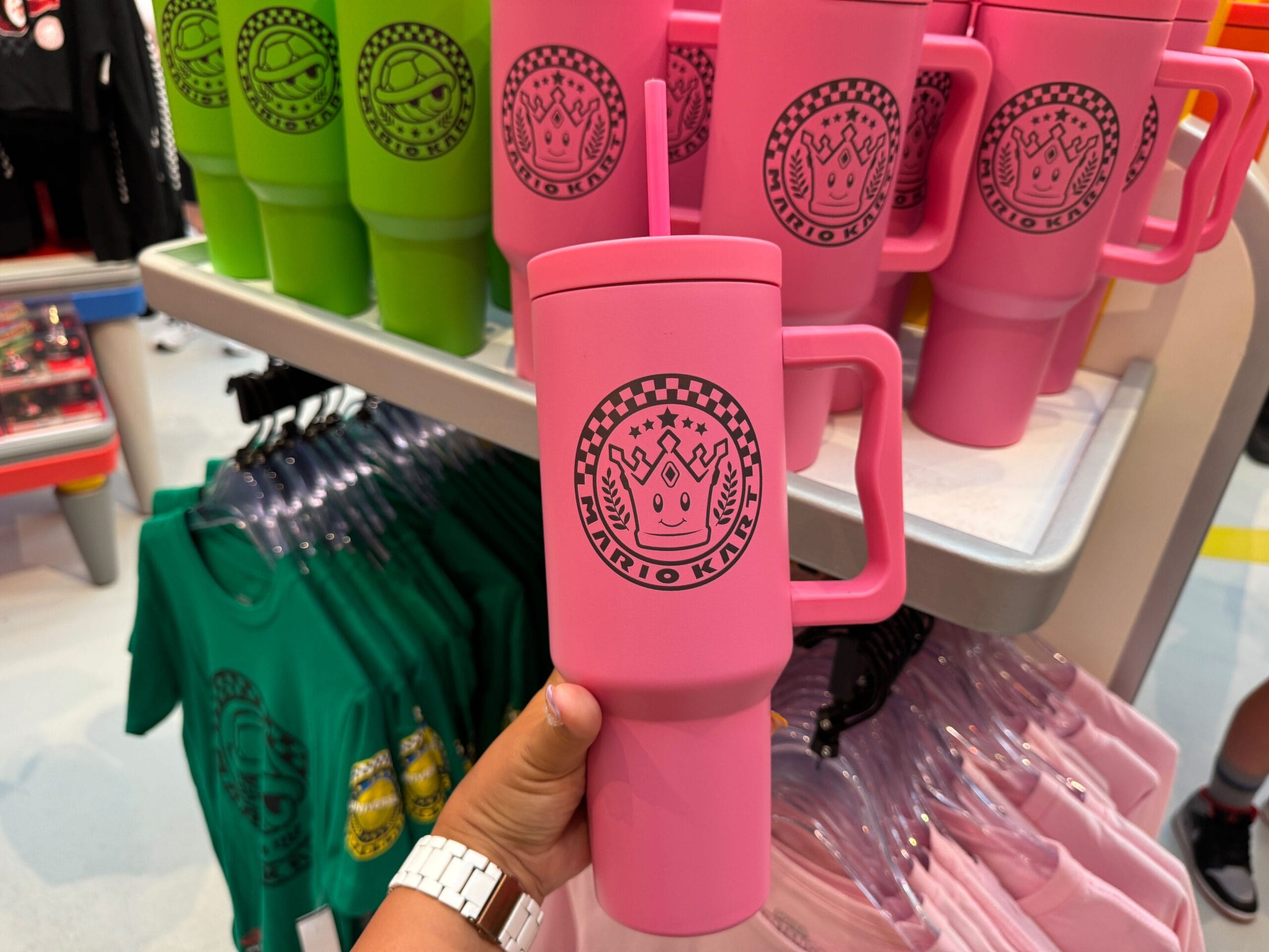 A hand holds a pink Mario Kart mug with a crown logo, Super Nintendo World merchandise—not a castle like those at Disney Parks.