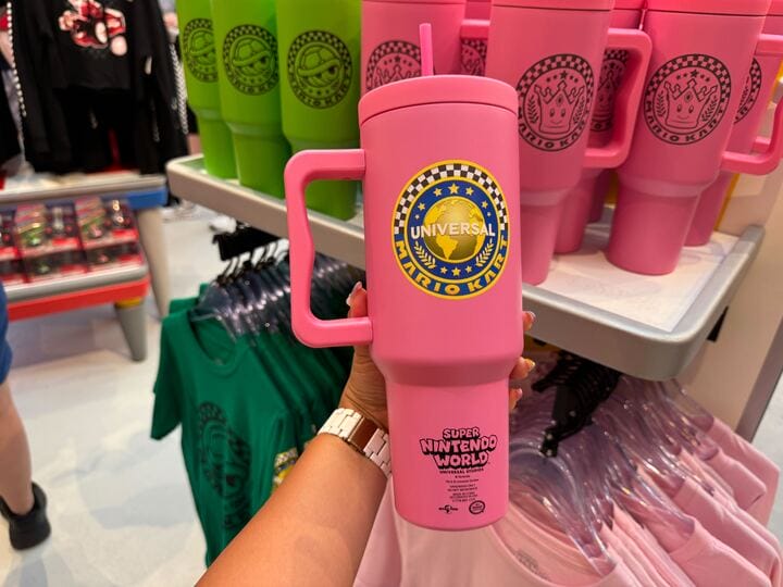 A hand holds a large pink Super Nintendo World mug among Mario Motors merchandise, a scene reminiscent of Disney park gift shops.