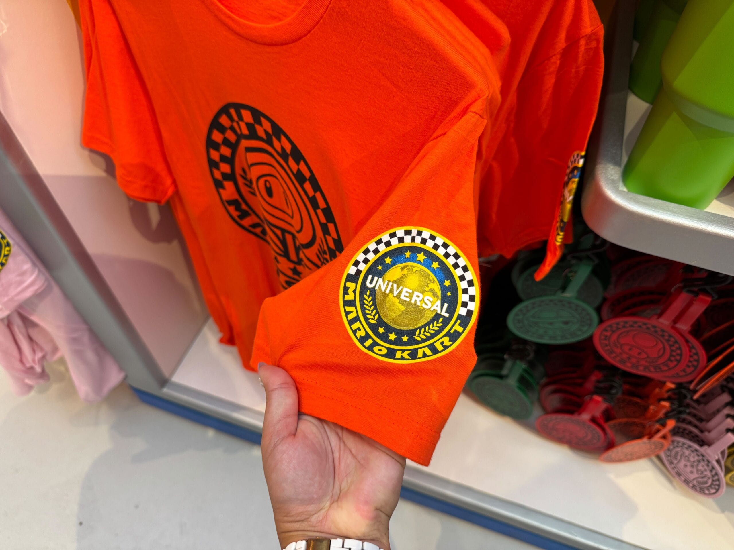 Hand holding an orange Mario Kart T-shirt with a Universal patch—distinctly not Disney, unlike castle merchandise at Disneyland.