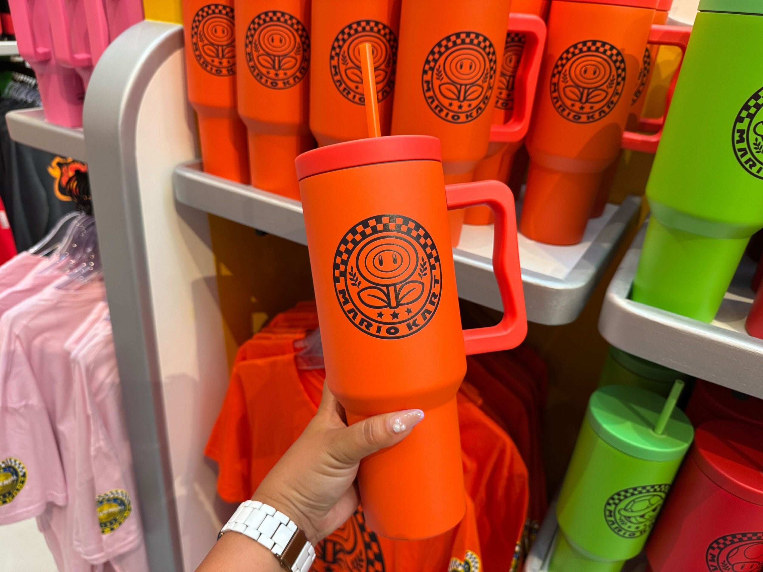 A hand holds an orange Mario Kart tumbler with a straw among themed merchandise and colorful shirts, reminiscent of Disney Park shops.