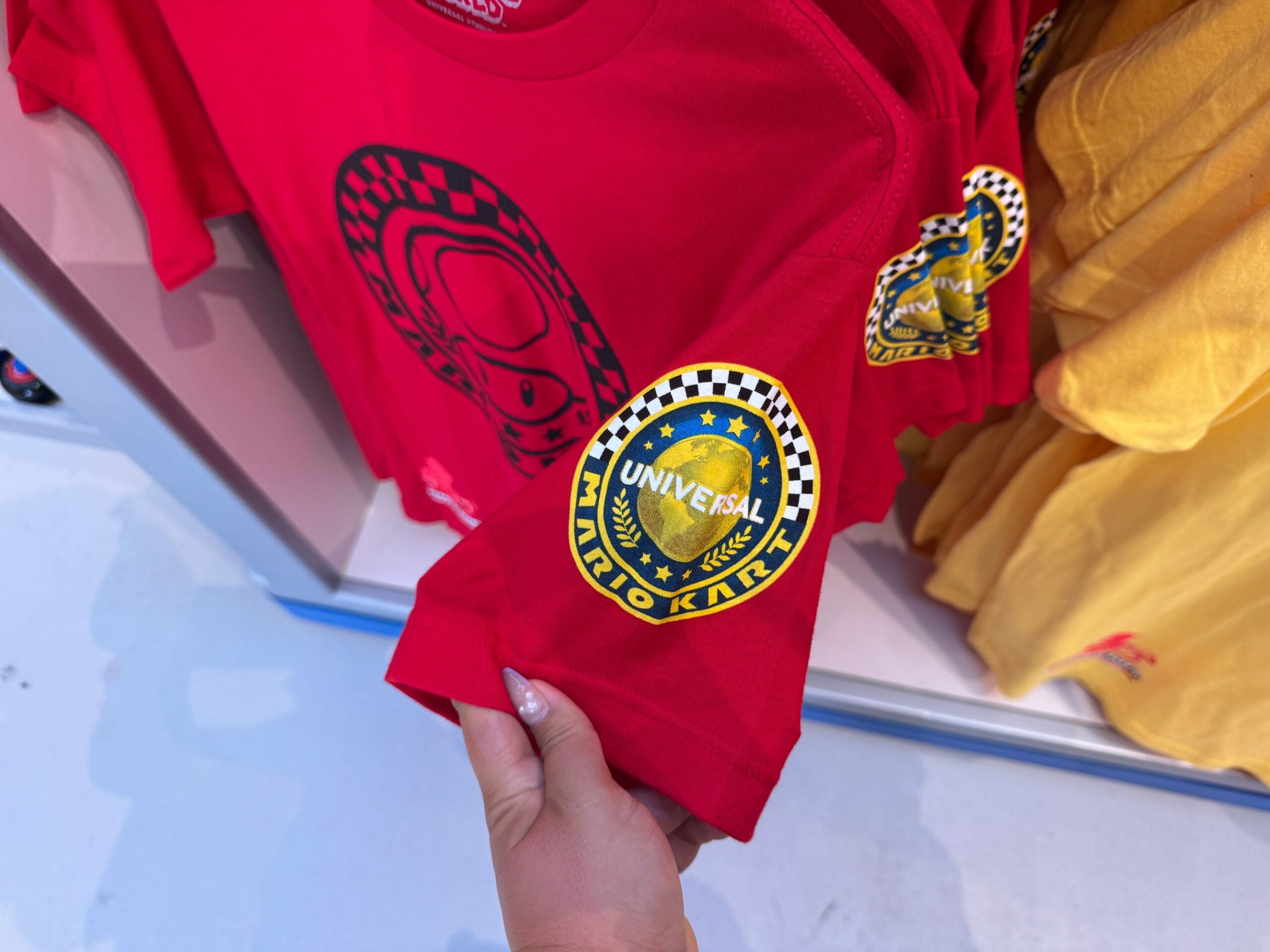 A hand holds a red Mario Kart t-shirt at Super Nintendo World—unlike Disney park shirts; yellow shirts hang beside, no castle visible.