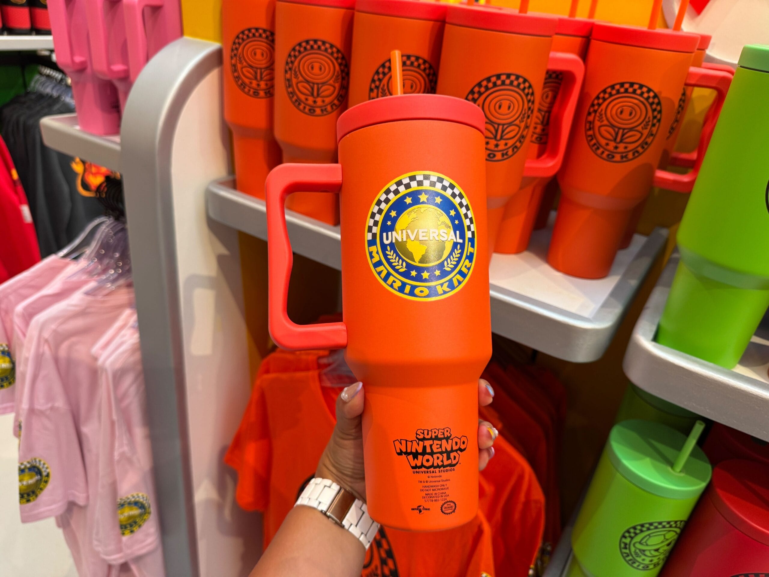 A hand holds an orange Mario Kart mug among Mario Motors merchandise, distinctly themed unlike any found in Disney Parks shops.