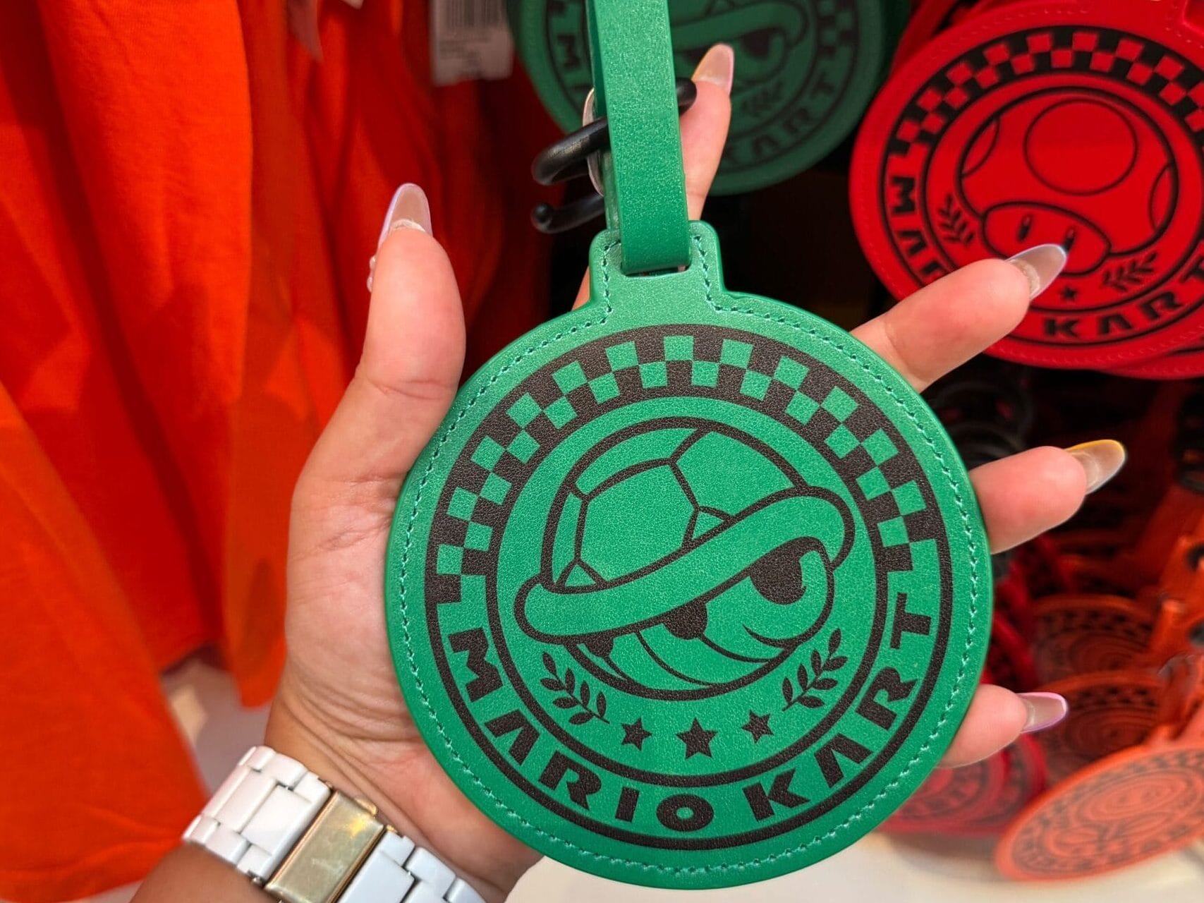 A hand holds a green Mario Motors luggage tag with a turtle shell design in a themed shop, similar to stores at Disney Parks.