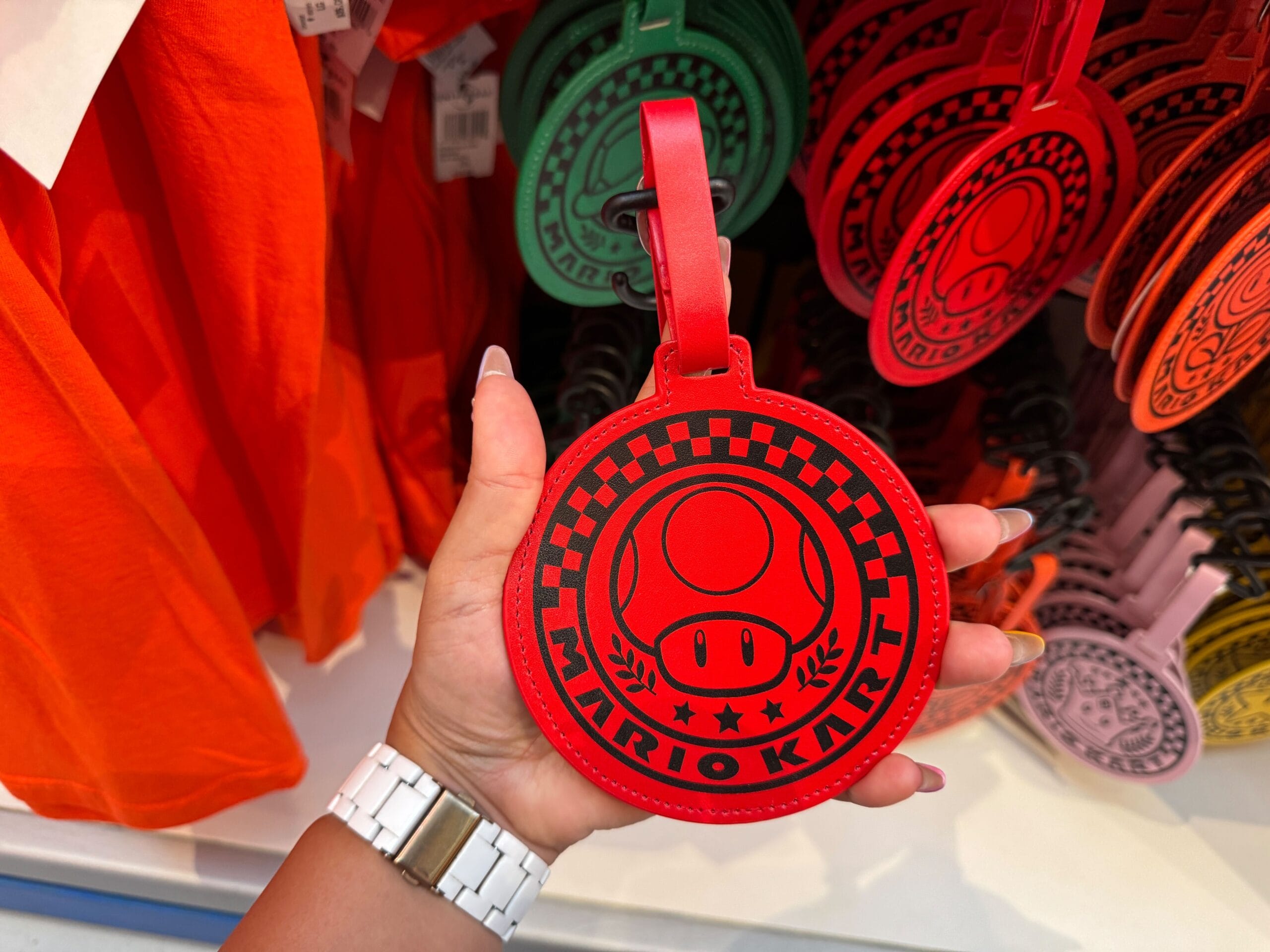 A hand holds a red Mario Kart luggage tag with a mushroom design, exclusive Super Nintendo World merchandise—not Disney Park merch.