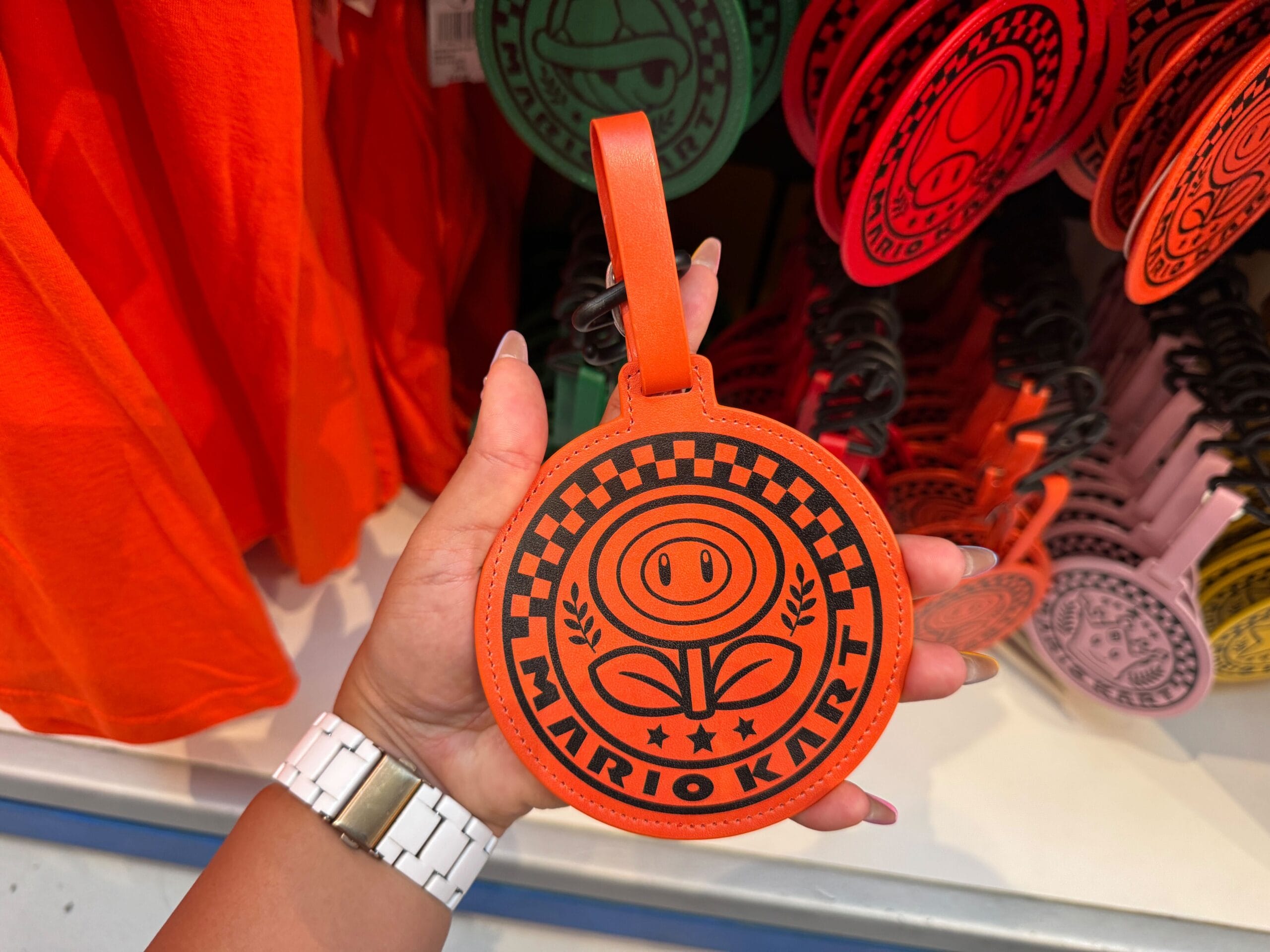 A hand holds an orange Mario Kart tag with a Fire Flower emblem, a fun nod to character merch like those found in Disney Parks stores.