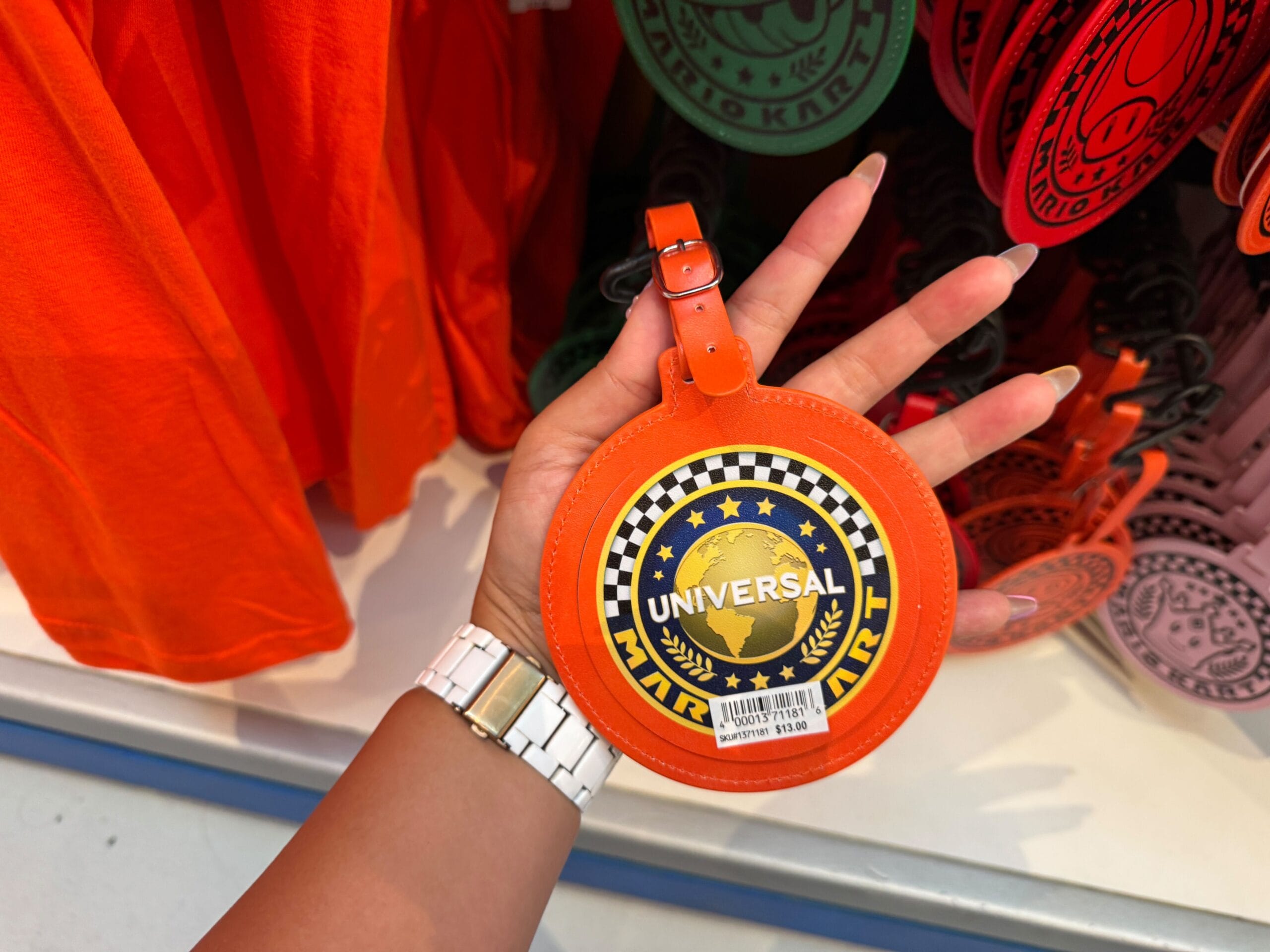 A hand holds an orange "Universal" Super Nintendo World Mario Motors luggage tag near orange and pink shirts, not Disney Parks merch.