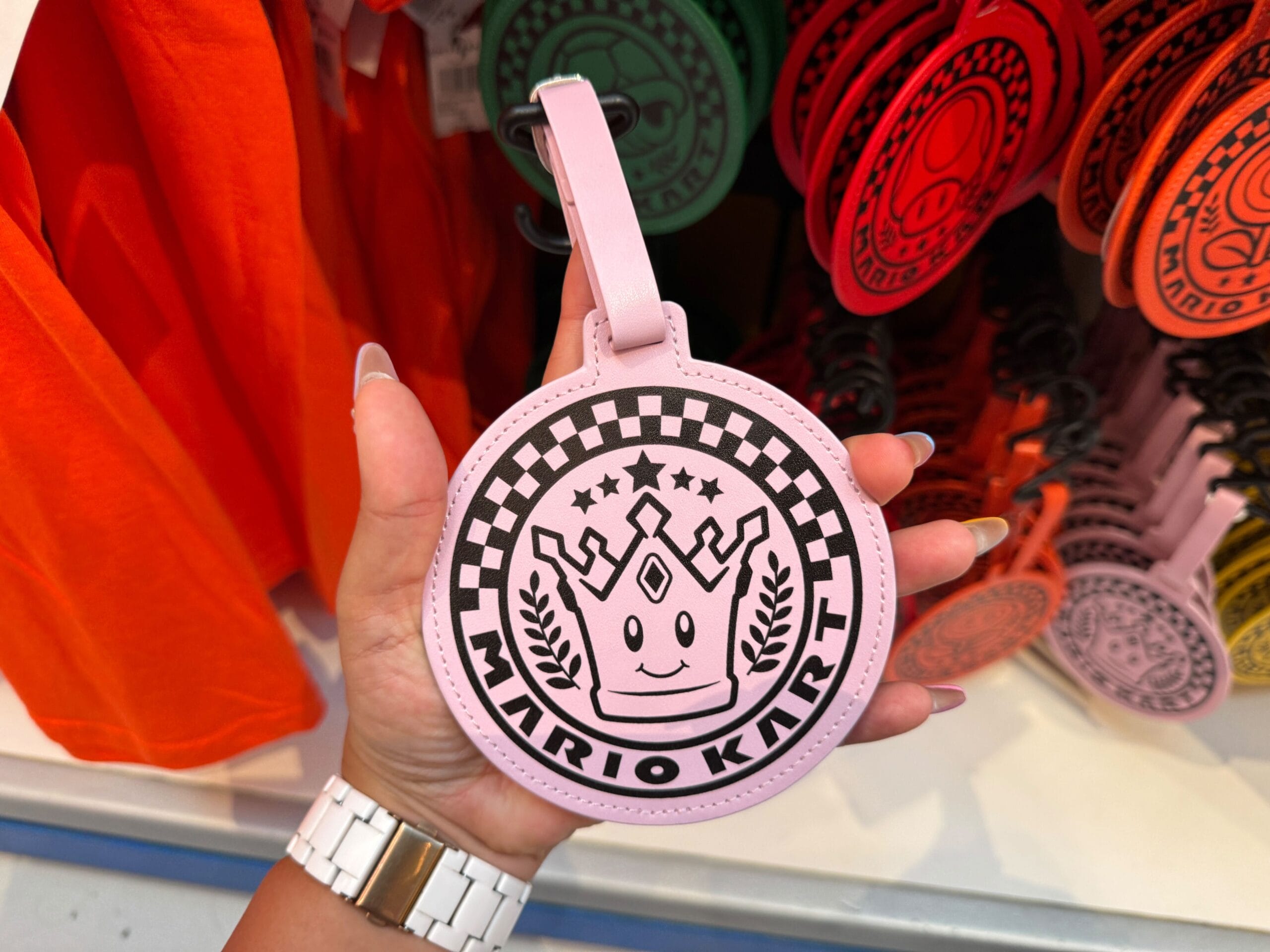 A hand holds a round, pink Mario Kart luggage tag with a crown and laurels—unlike any Walt Disney Parks castle merchandise.