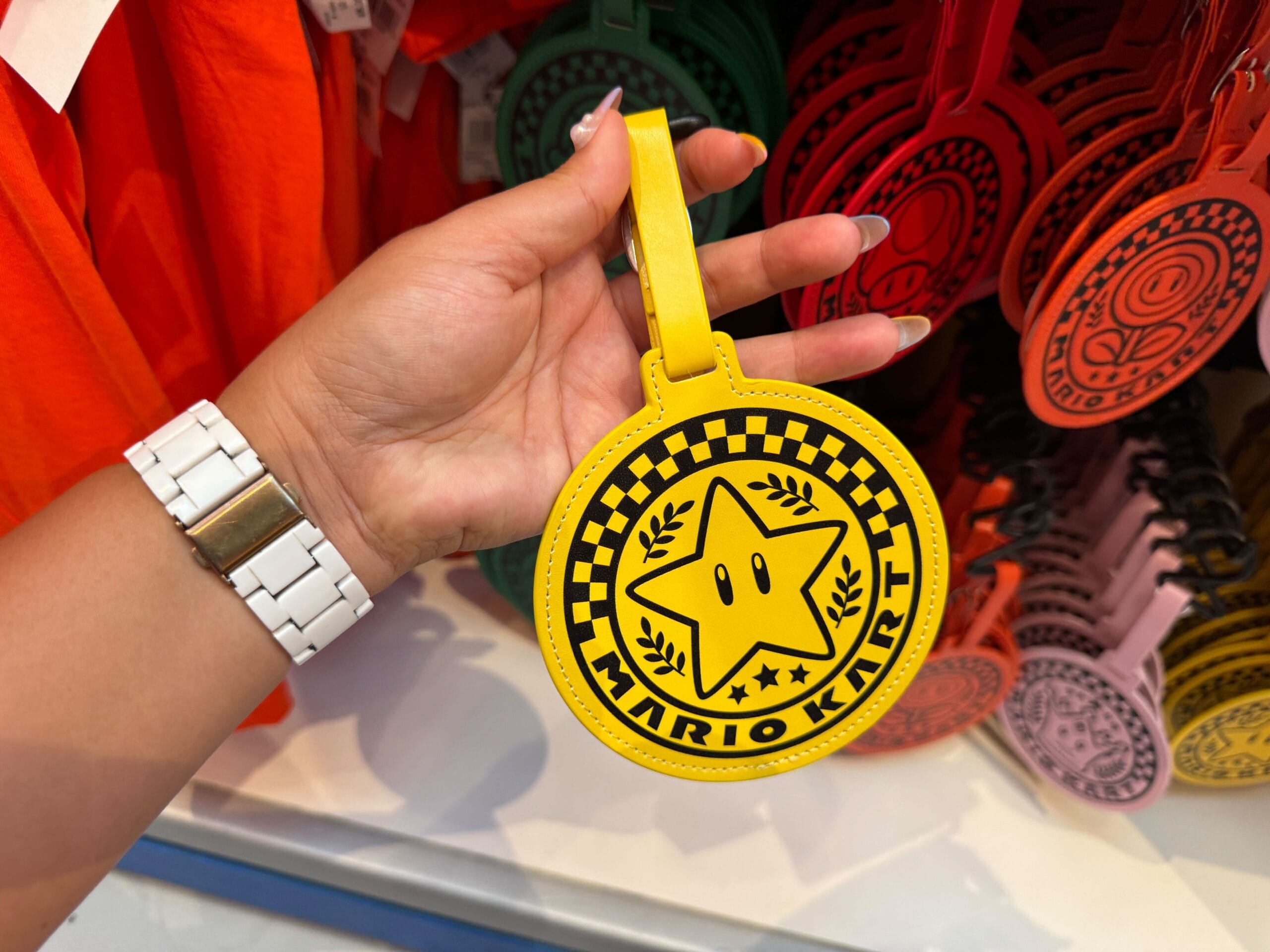 A hand holds a yellow Mario Kart merchandise luggage tag with a star and checkerboard pattern, similar to fun Disney parks souvenirs.