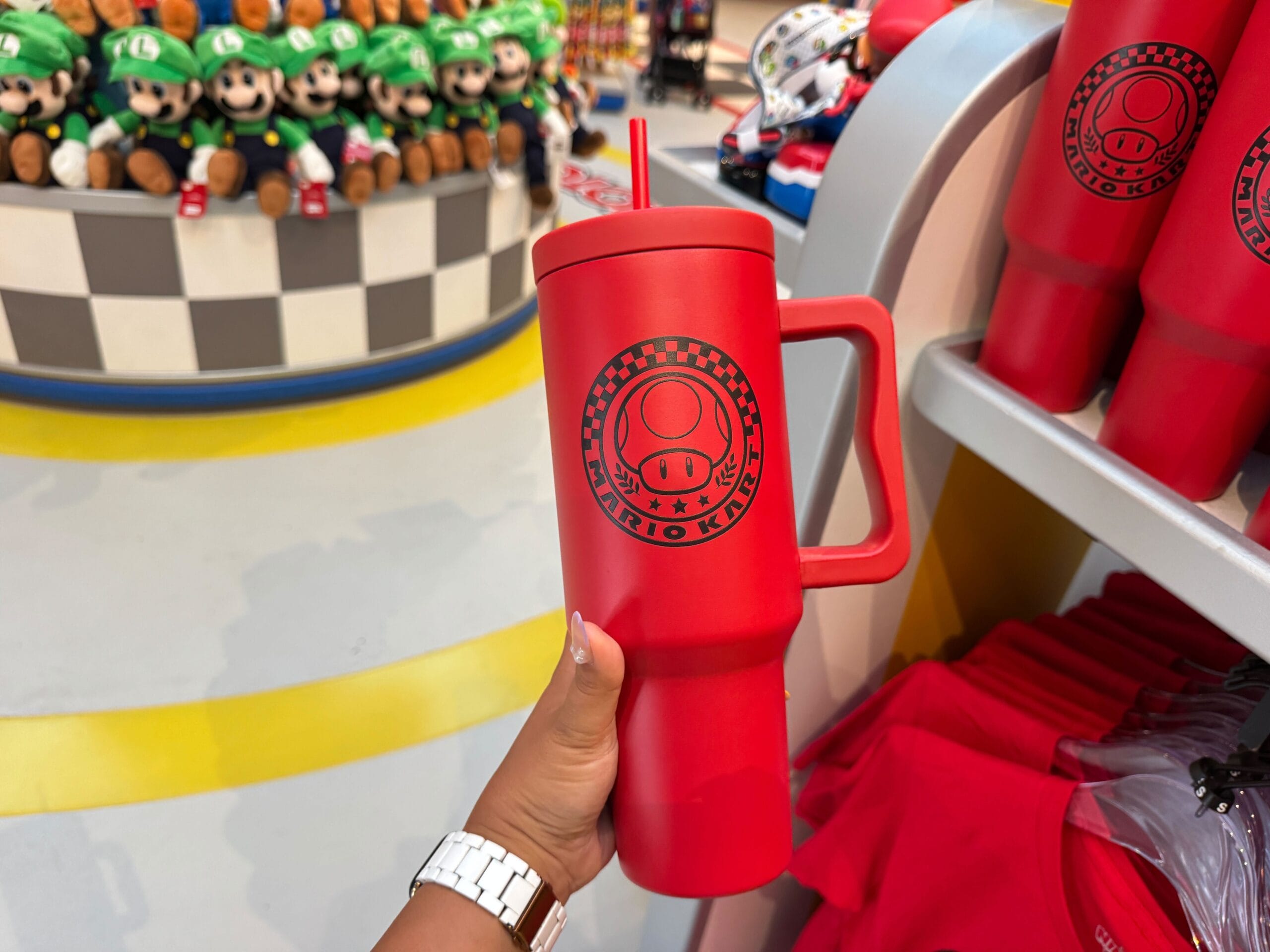 A hand holds a red Mario Kart cup with a straw, surrounded by plush toys—unlike Disney’s castle cups found in their parks.
