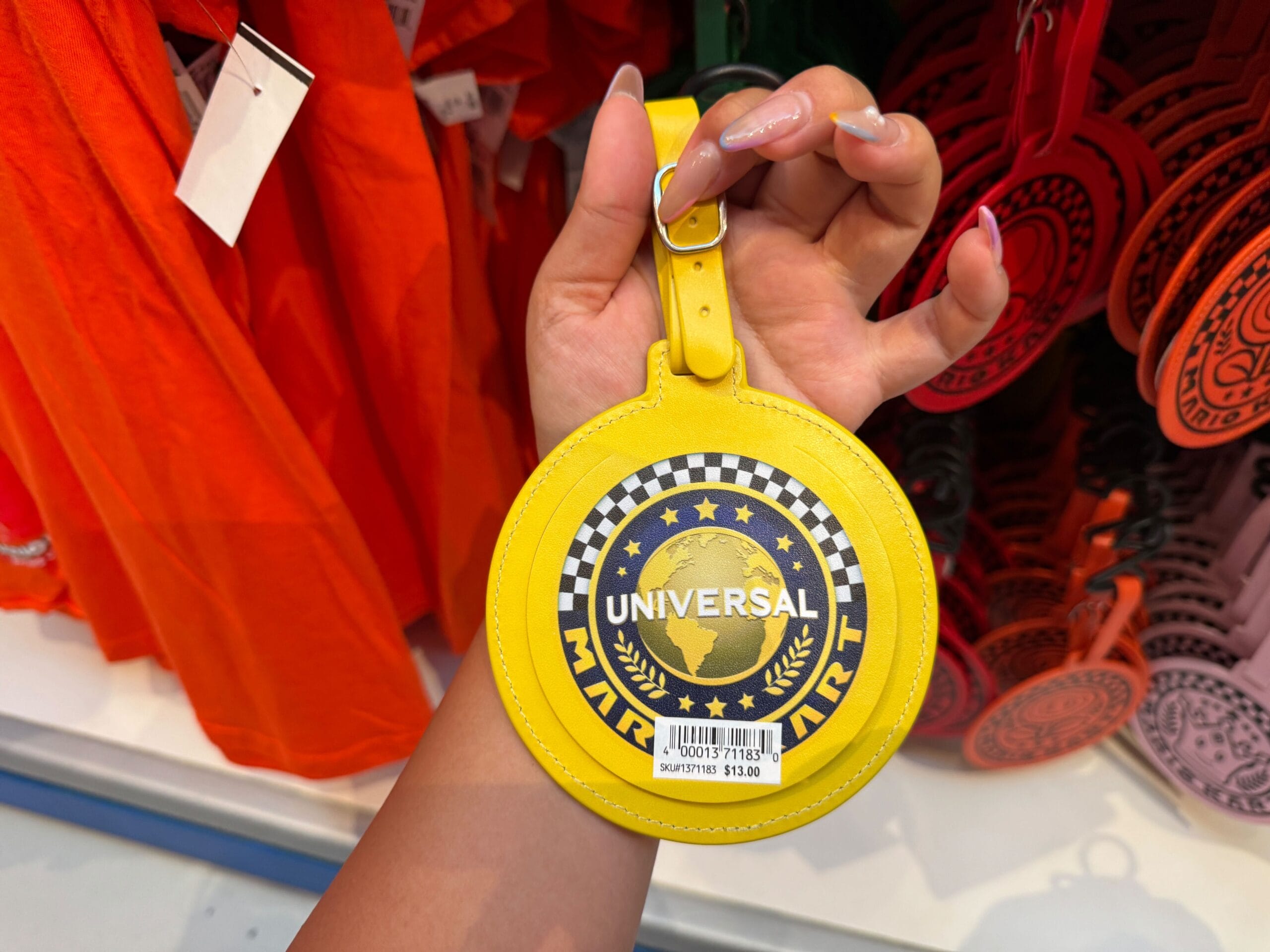 A hand holds a yellow Mario Kart luggage tag with a $13 sticker, displayed in a Universal shop, not at a Disney Park or castle.