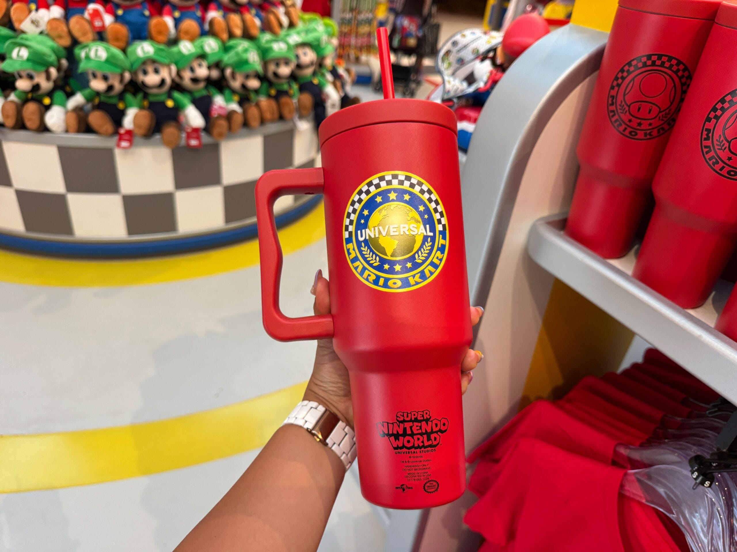 A hand holds a red Mario Kart cup from Super Nintendo World, surrounded by themed merchandise, similar to Disney park souvenir displays.