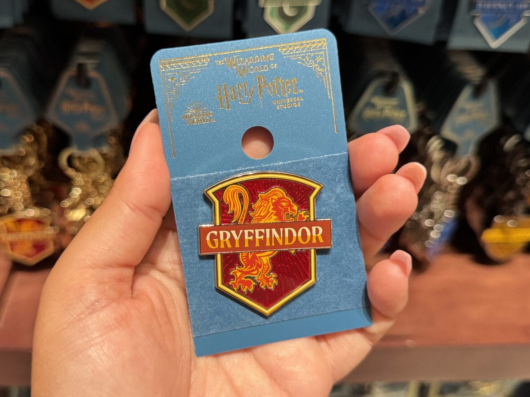A hand holds a Gryffindor badge in a store, surrounded by Wizarding World merchandise and house pins on display.