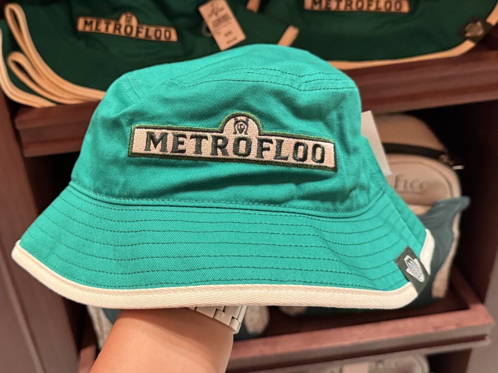 A hand holds a green "METROFLOO" bucket hat; more hats, perfect for Tour En Floo or Epic Universe trips, are in the background.