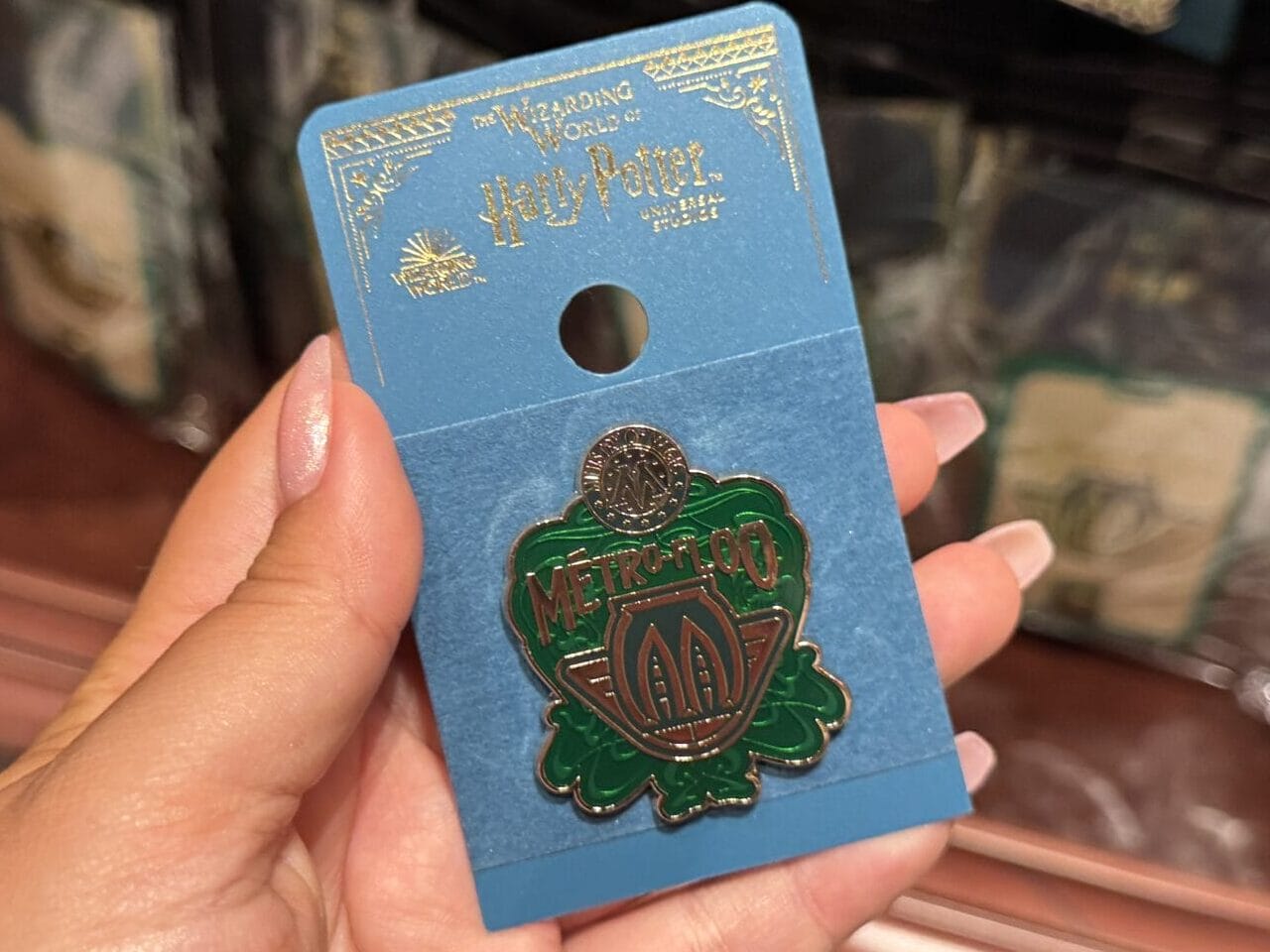 A hand holds a green Medusa enamel pin on a blue card—Epic Universe tour merchandise displayed in the background.