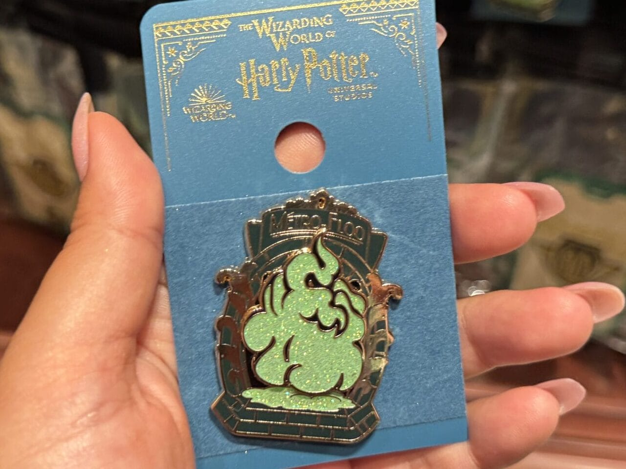 A hand holds The Wizarding World of Harry Potter merchandise at an Epic Universe Slytherin enamel pin display.