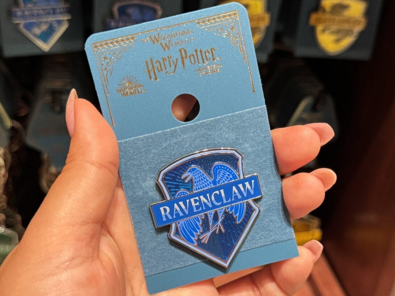 A hand holds a blue Harry Potter Ravenclaw pin—magical Harry Potter merchandise on display at Epic Universe.