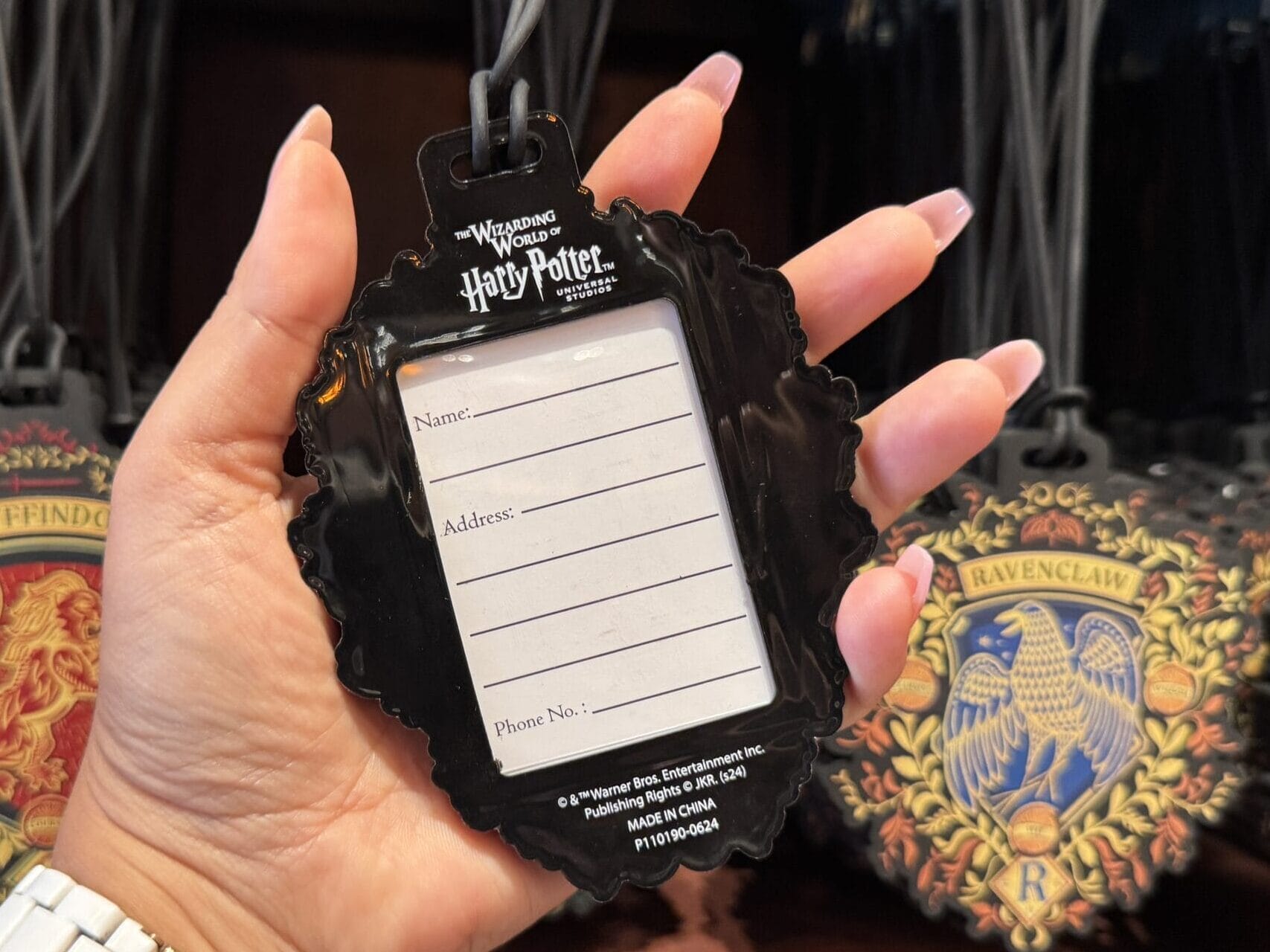 Hand holds a Wizarding World of Harry Potter merchandise tag; house tags in background—perfect for your next Epic Universe tour en floo.