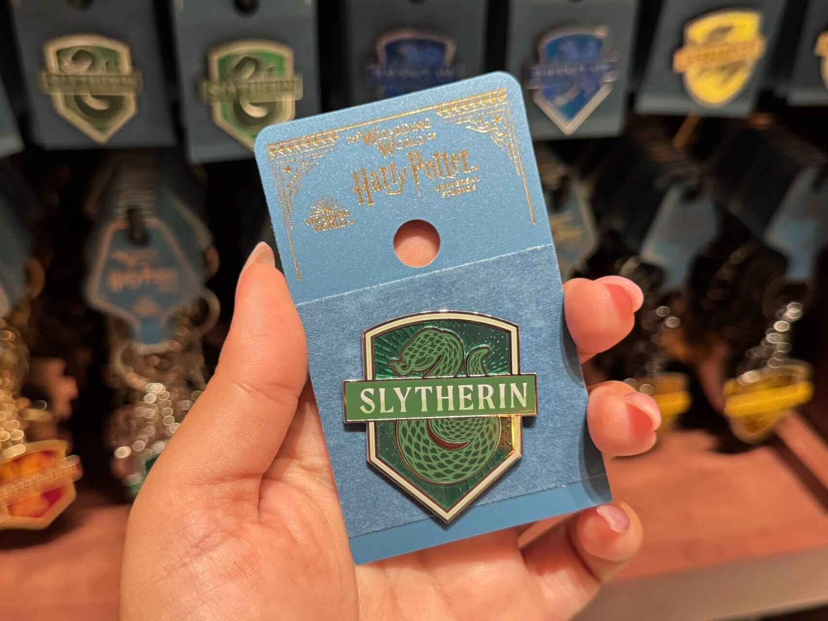 A hand holds a Slytherin pin from Harry Potter among Wizarding World pins on an Epic Universe store display.