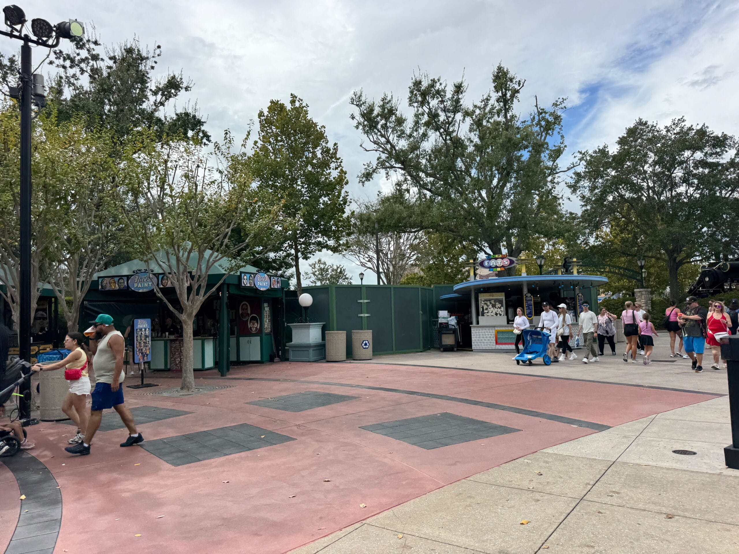 Guests stroll by shuttered snack stands and construction barriers, under trees and cloudy skies—very different from Disney parks.