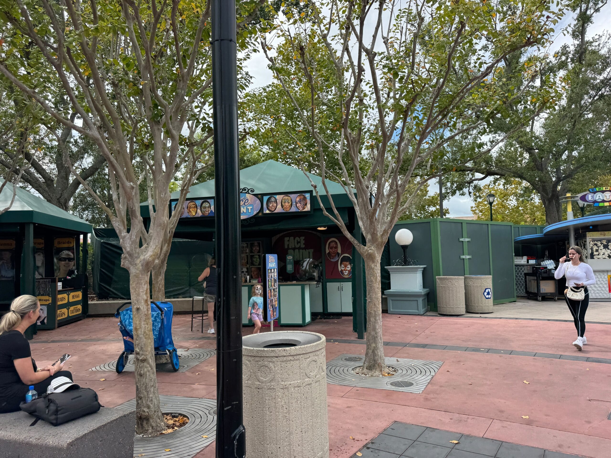 Outdoor caricature stand by guests, trees, and trash cans, with construction walls in a theme park area not resembling Disney castles.