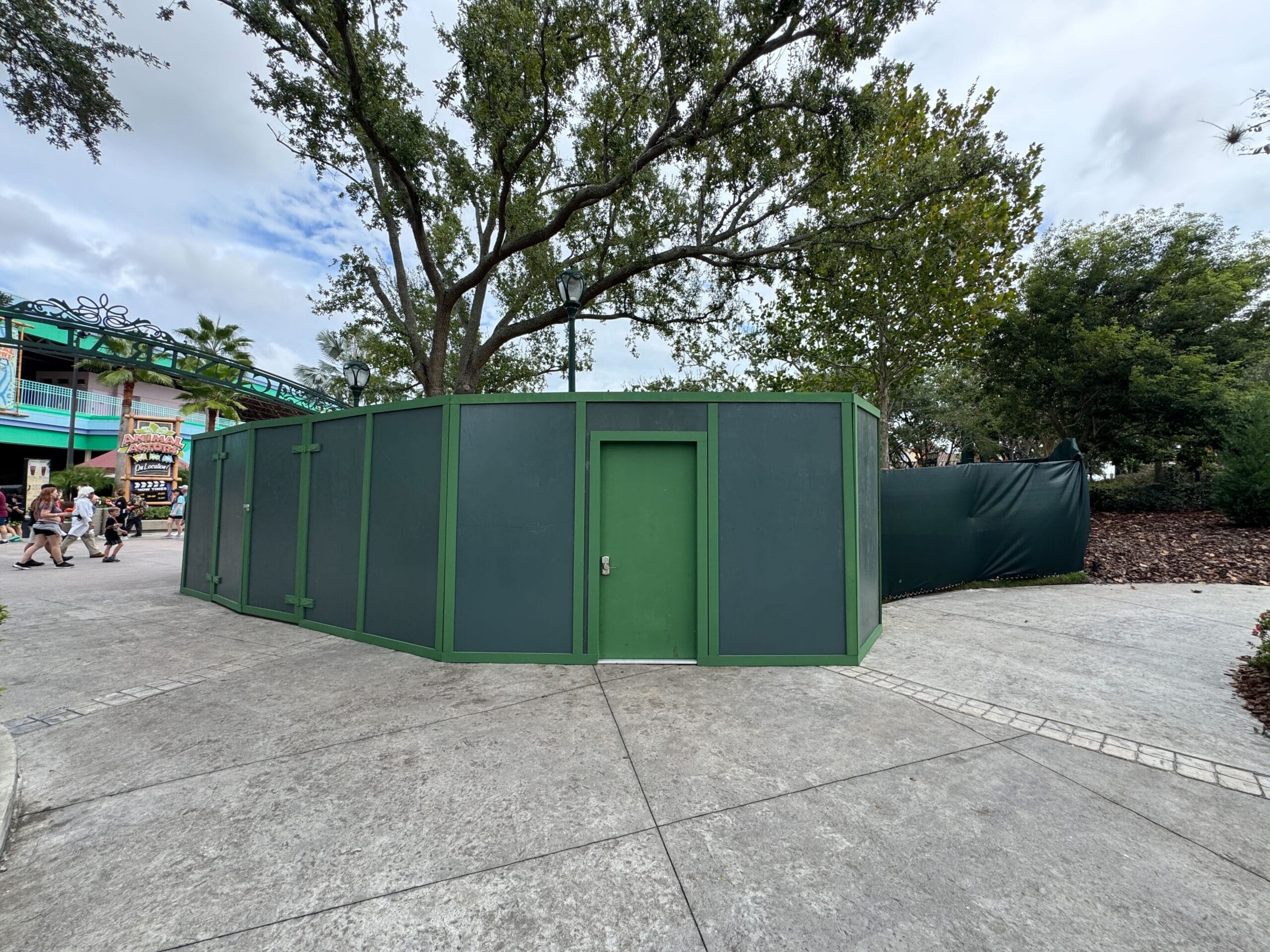 Green construction walls and a door block future food kiosk; feels like EPCOT renovations, trees and clouds overhead. No castle here.