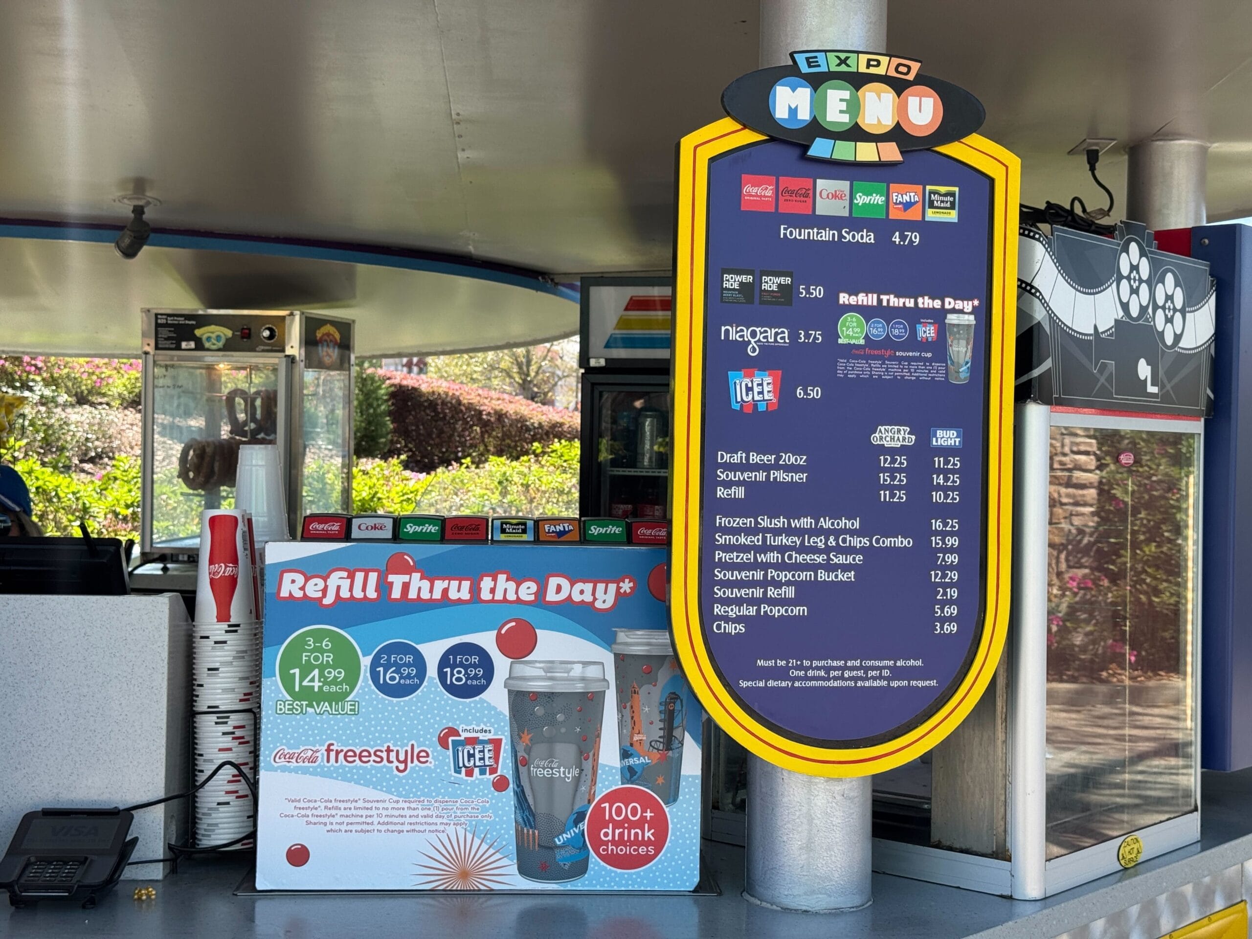 Outdoor food kiosk by construction walls with drink refill station and cup promo, similar to kiosks found in Disney parks.