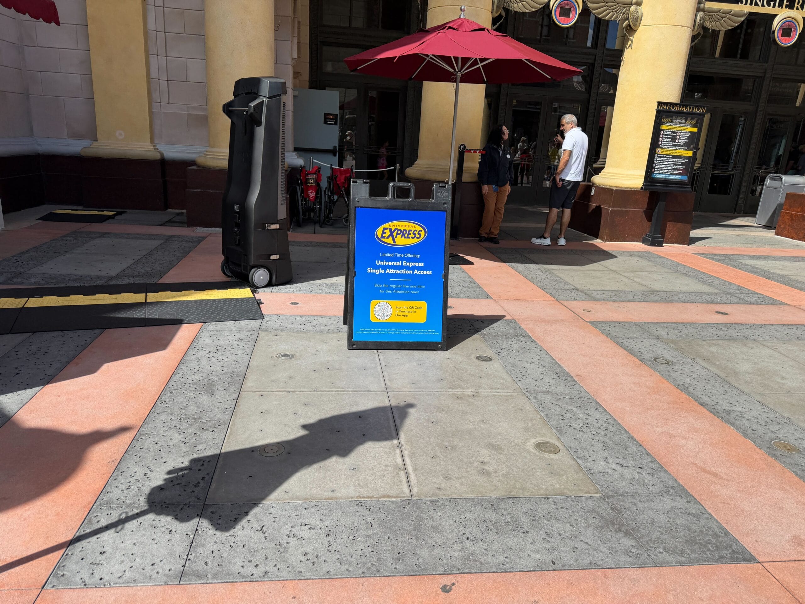 Blue Express Pass sign with yellow text offers Express Access near an entrance, crowds and umbrellas behind—not a Disney castle.