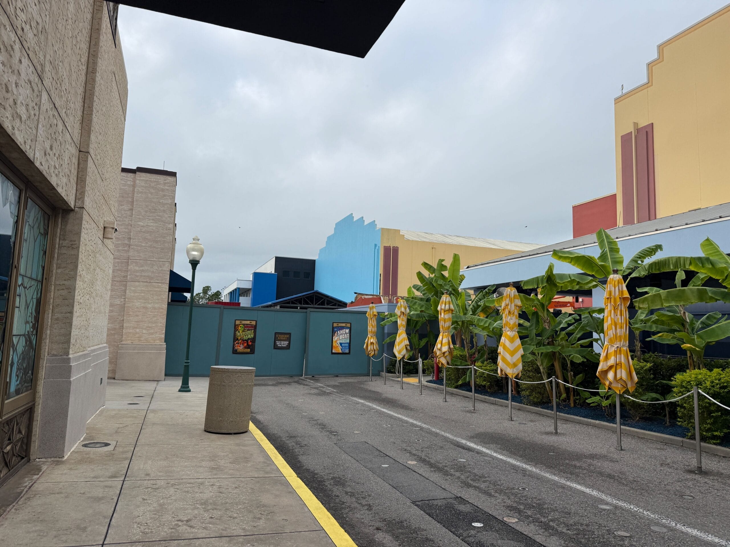 A quiet street with yellow-striped umbrellas, banana plants, and vibrant buildings—evoking Main Street vibes at a Disney park.