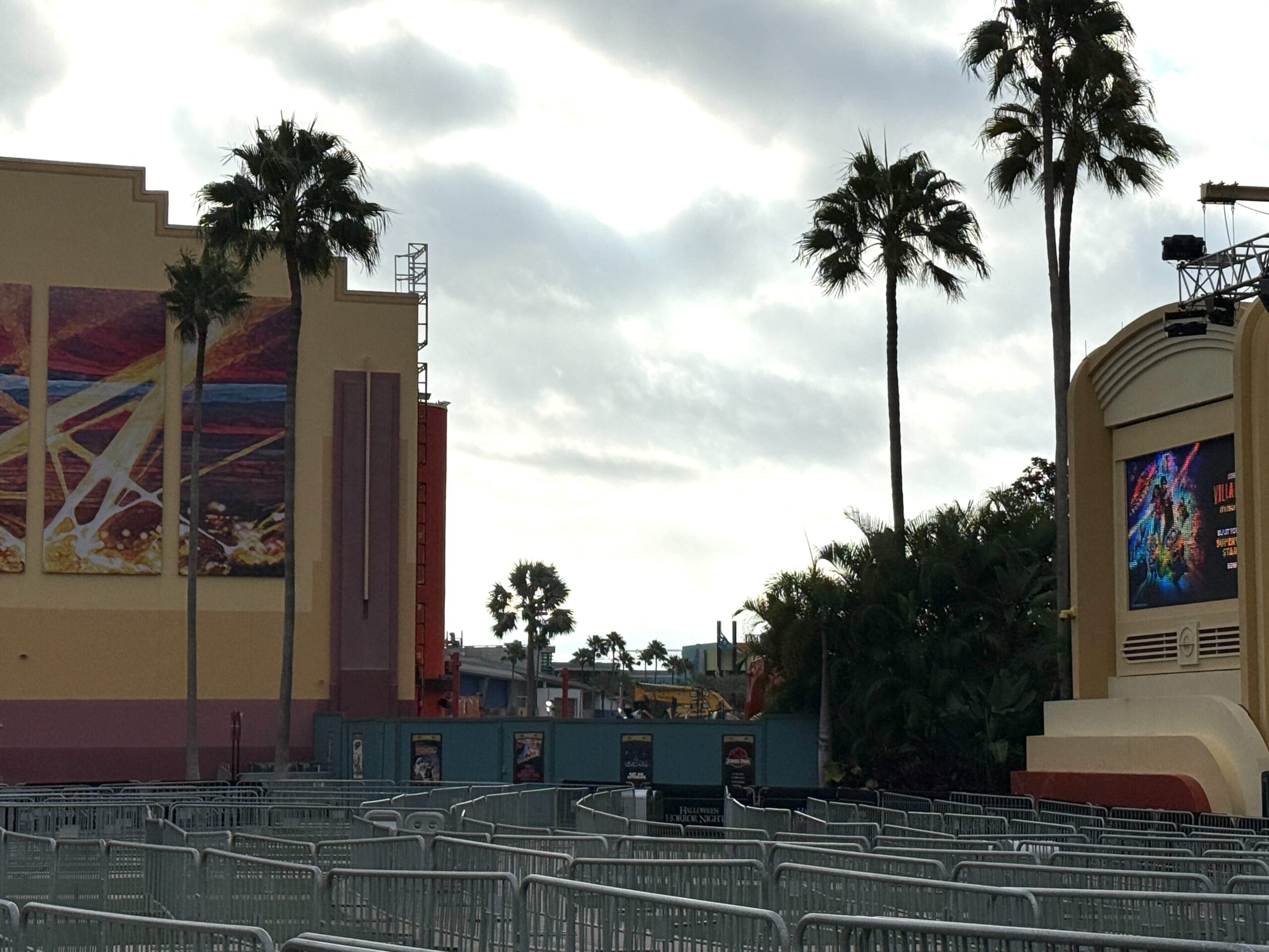 Outdoor queue with metal barriers, palm trees, murals, and a digital screen