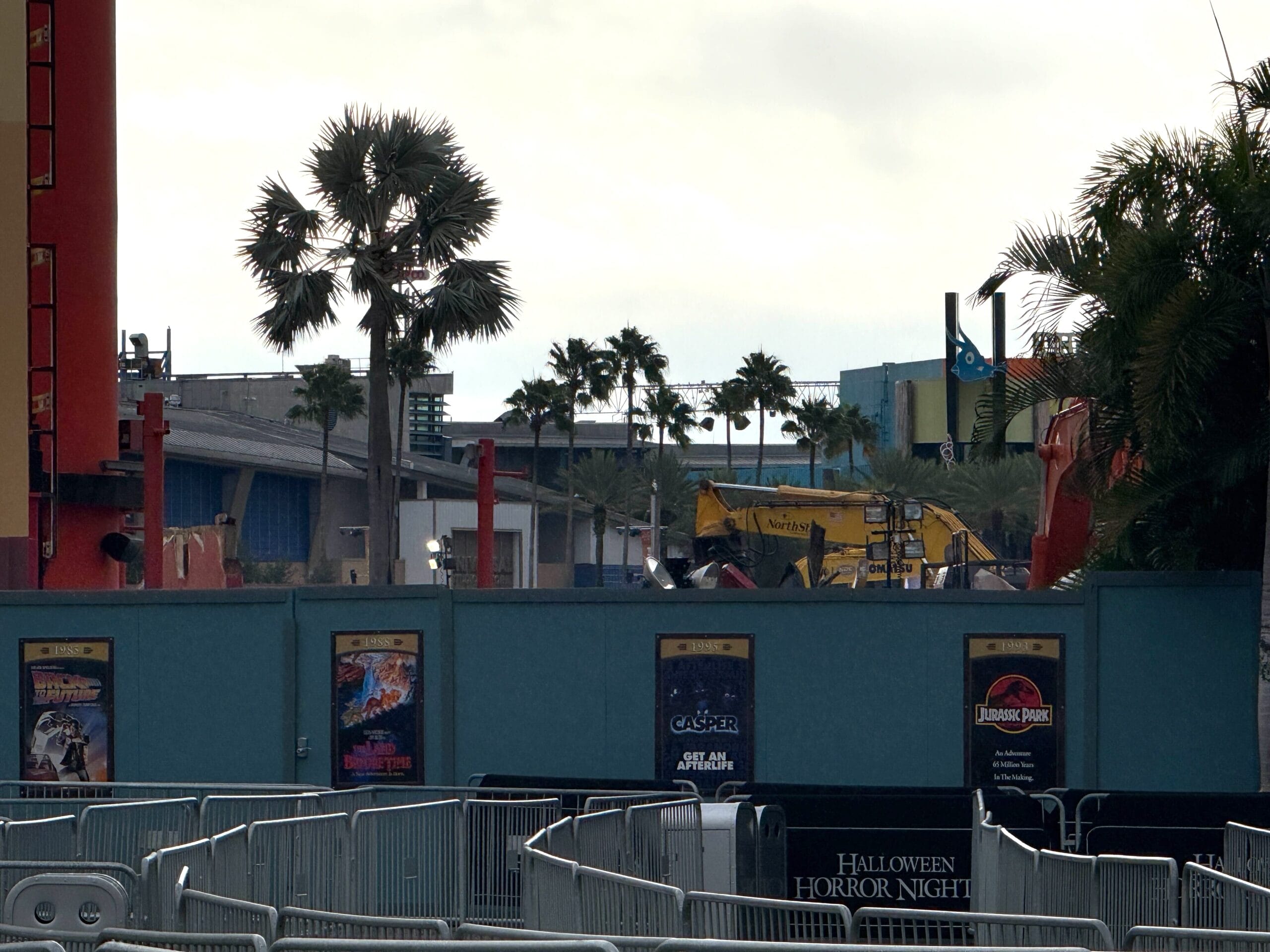 Construction site with palm trees, roller coaster tracks, and a blue fence and posters.