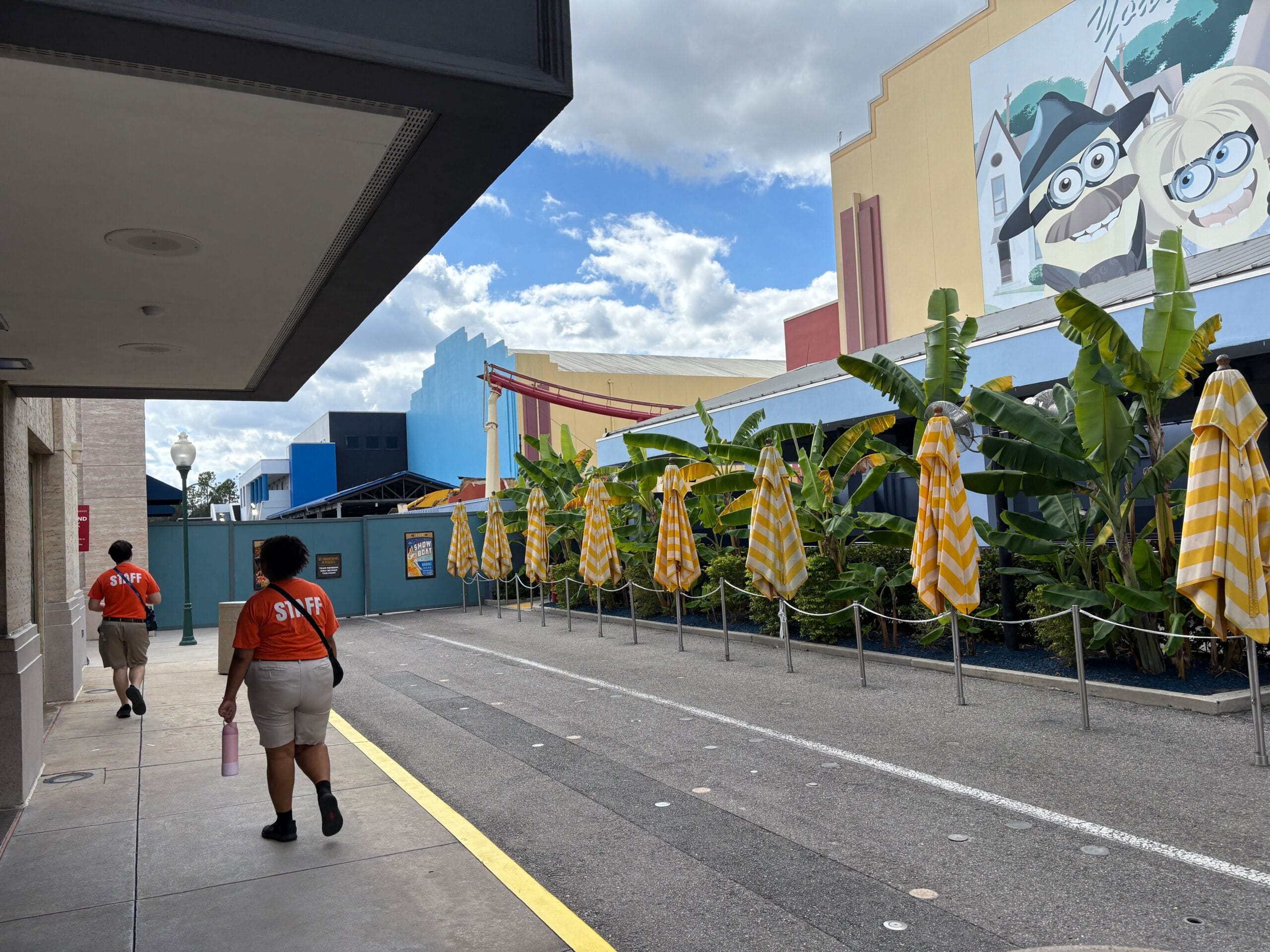 Two staff walk by yellow-striped umbrellas and Minions cartoon art near construction walls at Universal Studios Florida. A small portion of the remaining Rip Ride Rockit track is visible in the distance.