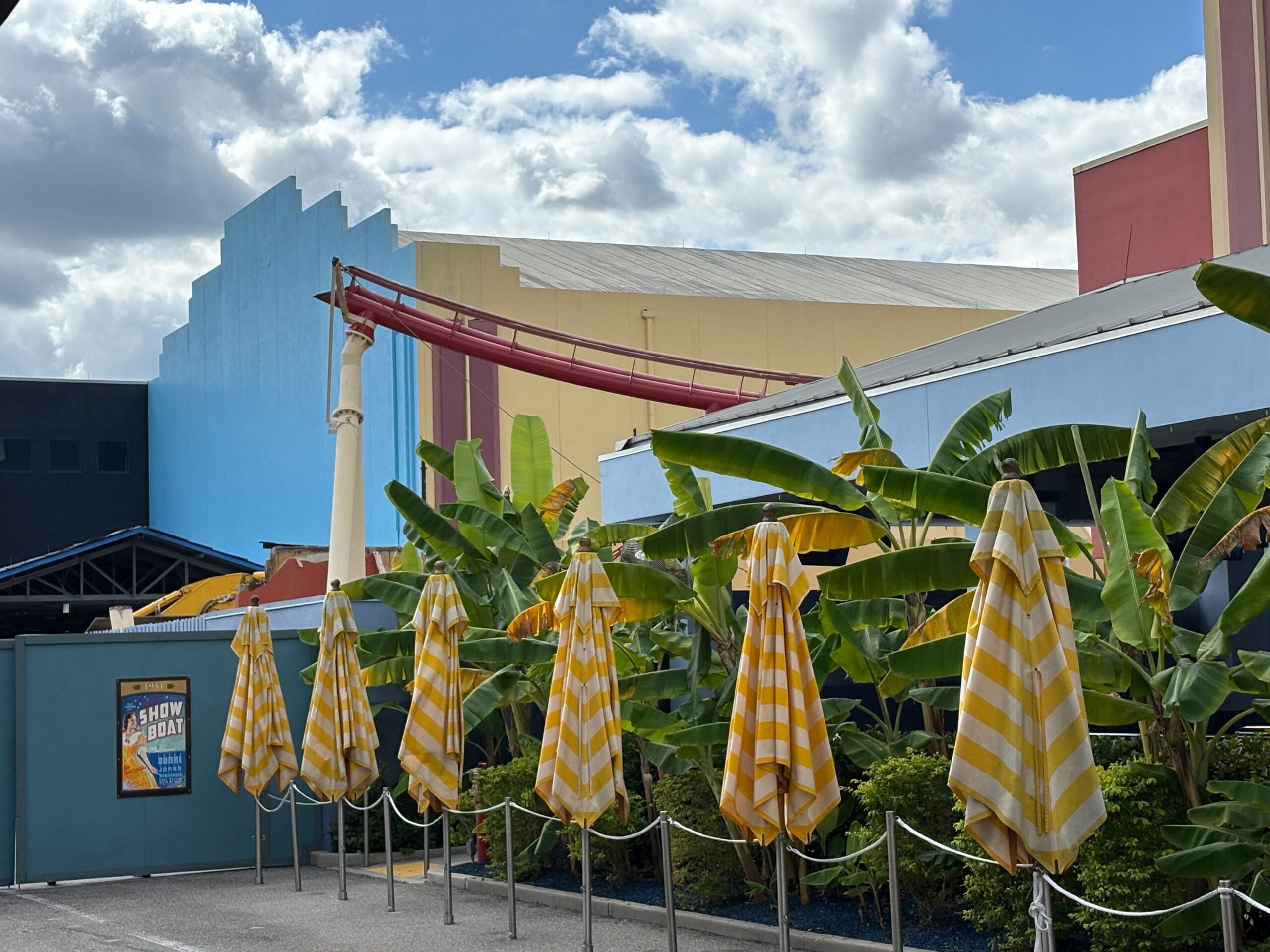 Closed yellow striped umbrellas stand by banana plants and construction walls at Universal Studios Florida with a small portion of the remaining Rip Ride Rockit track visible behind.