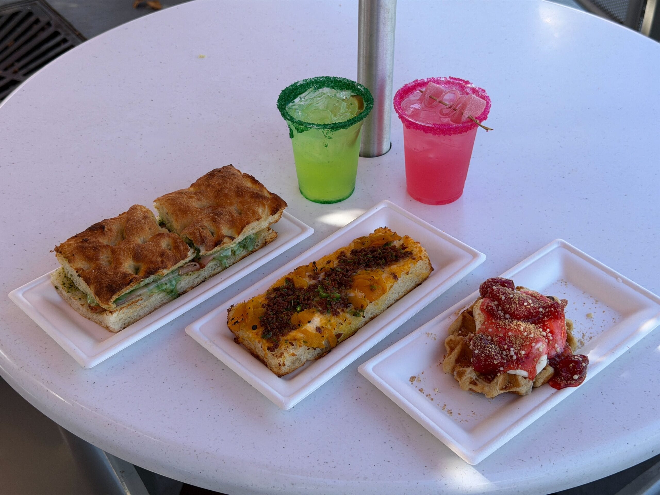Three white plates of Wicked food items and two vibrant drinks on an outdoor table, perfect for a magical snack break.
