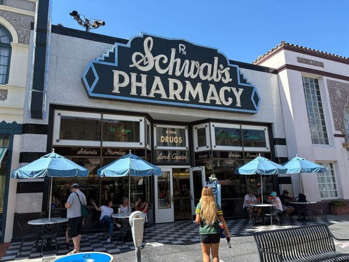 Guests relax under blue umbrellas outside Schwab's Pharmacy, a vintage spot in Universal Studios, not one of Disney's castle parks.
