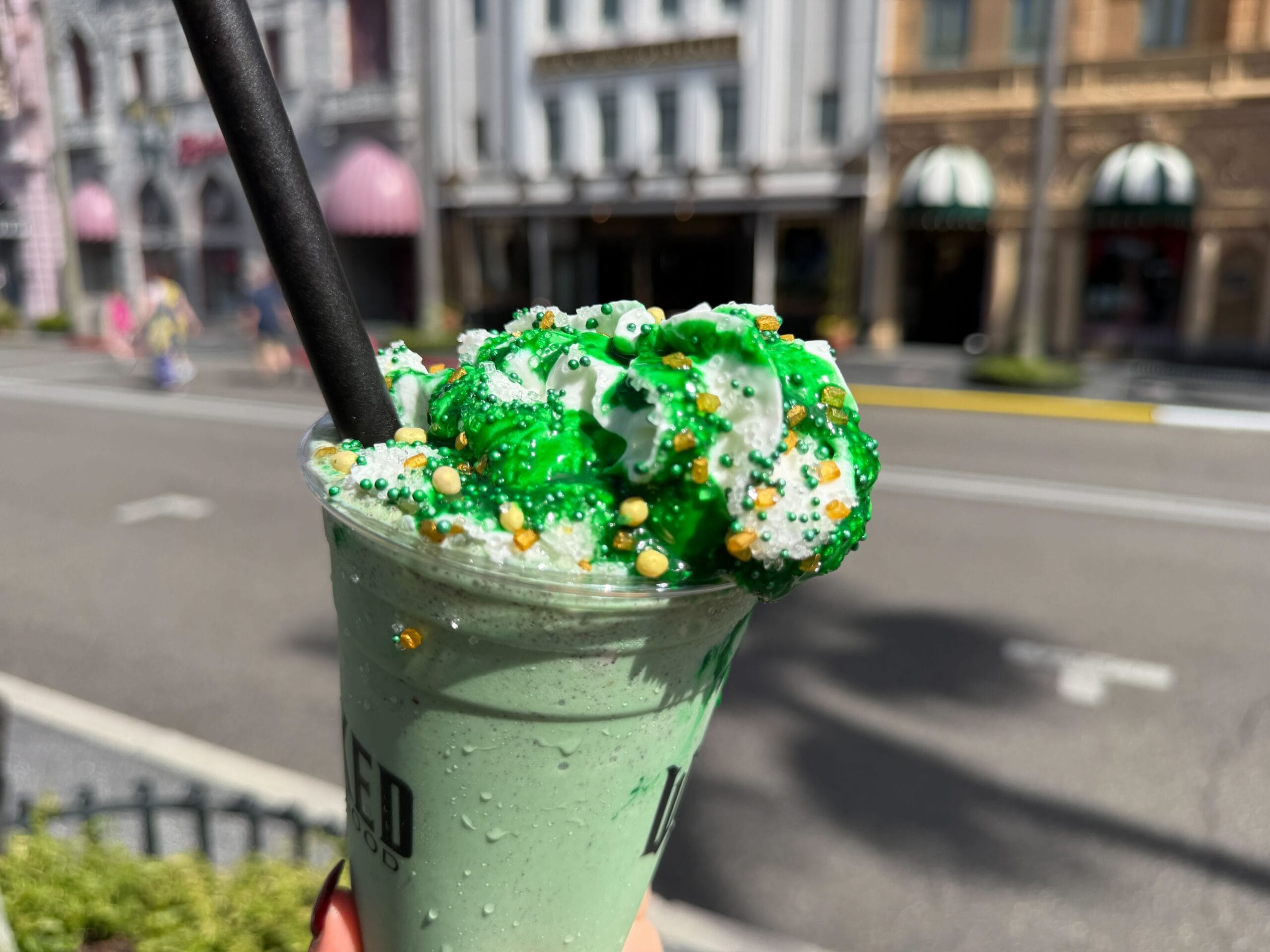 A green Wicked Drinks milkshake topped with whipped cream and sprinkles sits on a city street at Universal, not Disney Parks.