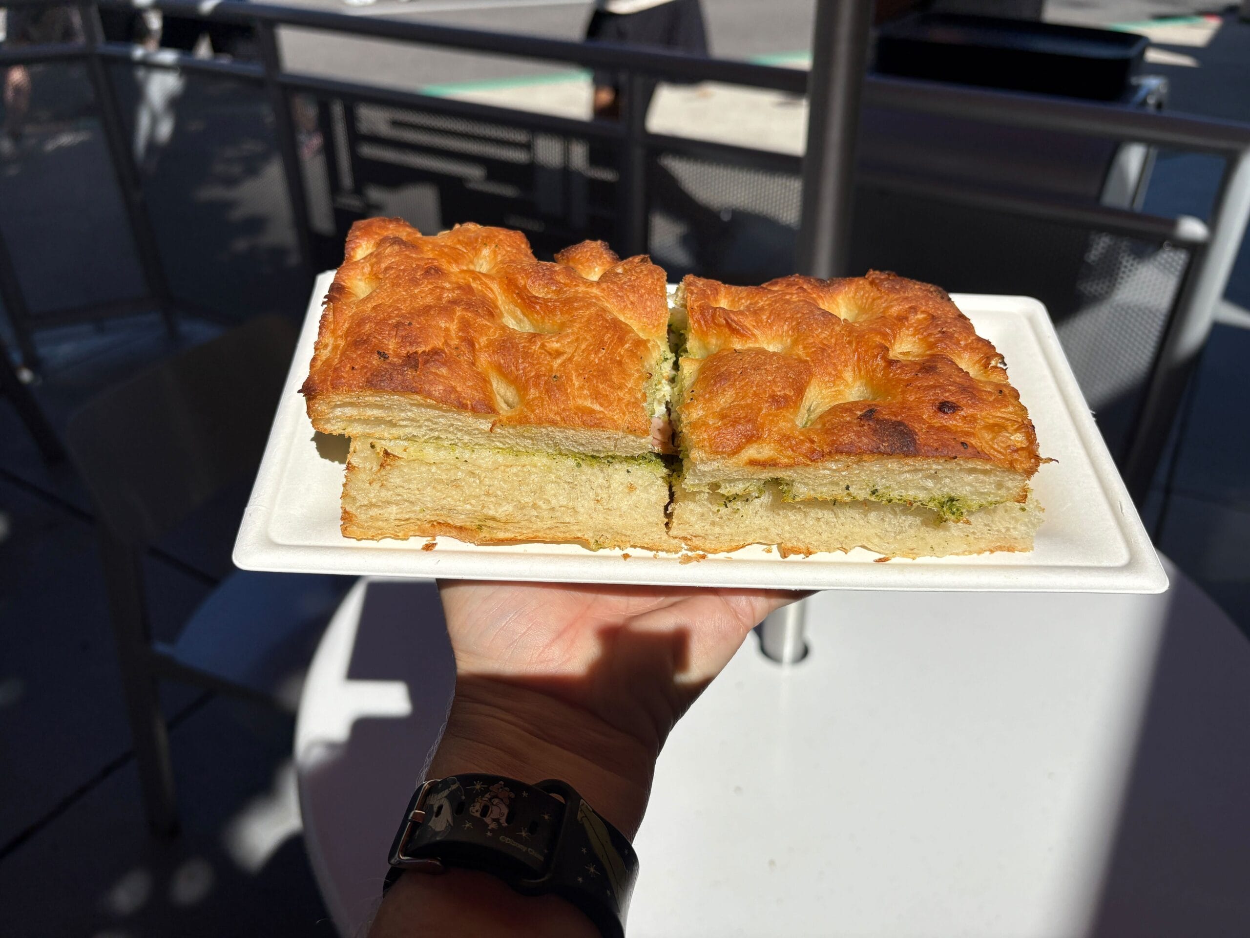 A hand holds a plate with two thick focaccia sandwiches in the sunshine, outdoors at Disney Park seating—no castle in view.