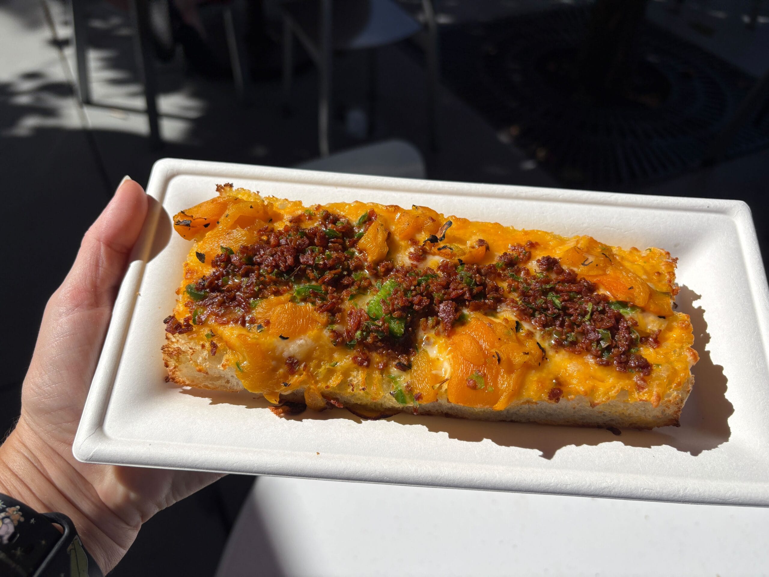 A hand holds a cheesy bread slice with herbs and crispy bits, served on a white tray outdoors at Universal Orlando.