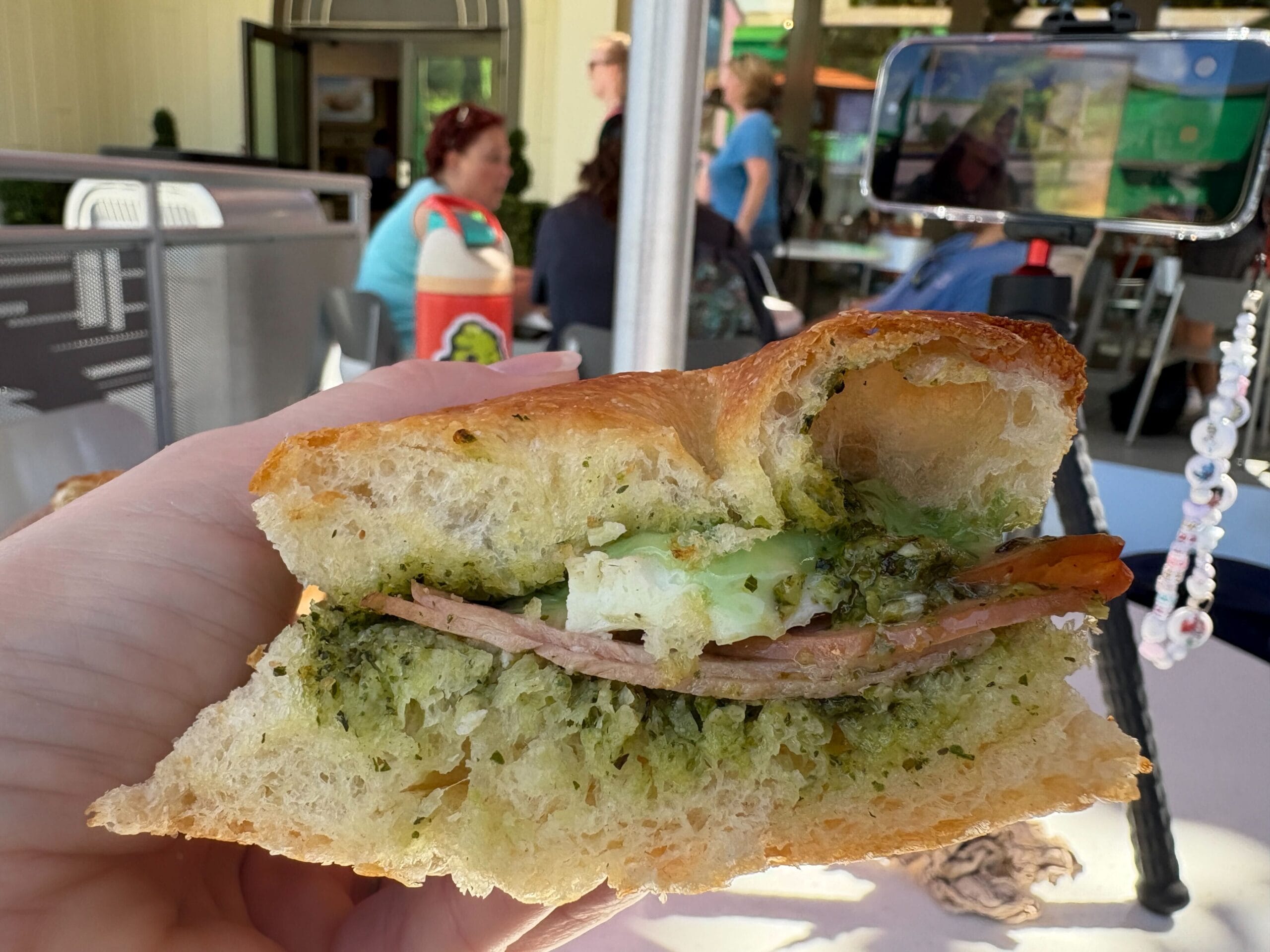 A hand holds a focaccia sandwich with pesto, ham, and mozzarella as people dine outdoors like at Epcot’s Italy Pavilion in Disney World.
