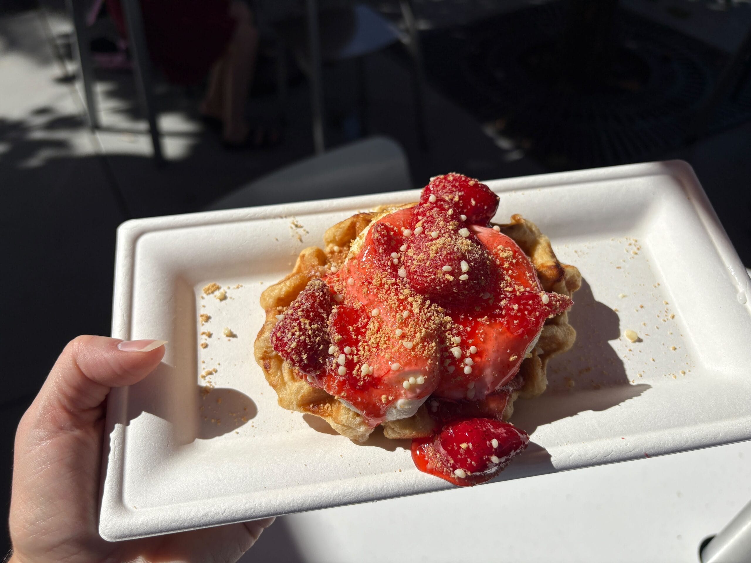 A hand holds a waffle topped with strawberries, cream, and crumbs on a white tray in sunlight, looking like a Disney park treat.