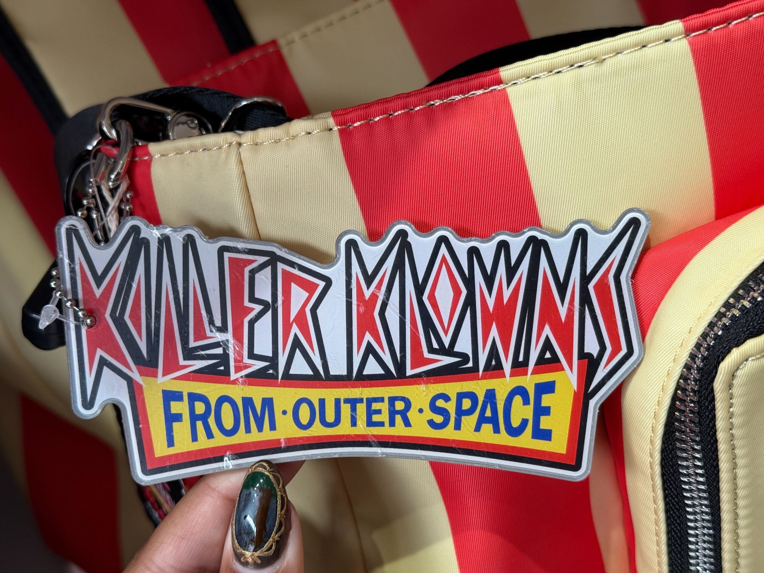A hand holds a "Killer Klowns from Outer Space" tag attached to a striped red and beige bag.
