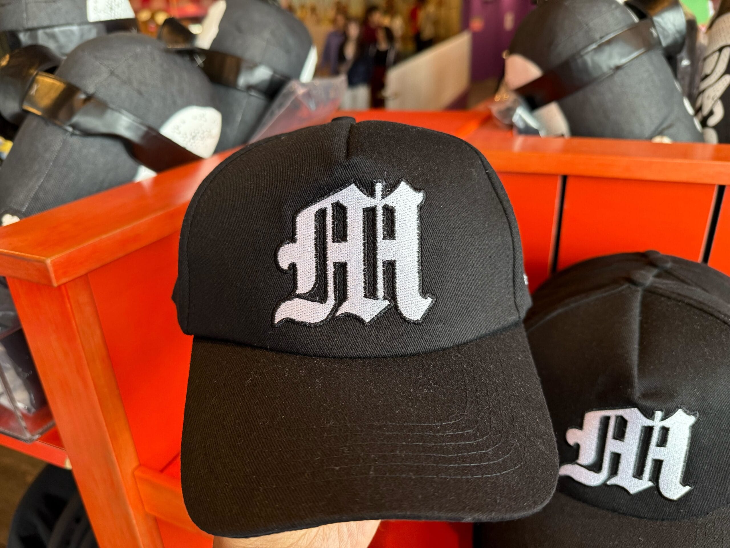 A black baseball cap with a white gothic "M" logo, Emo Minion Merchandise, displayed on a red shelf at Universal Studios—no Disney castle.