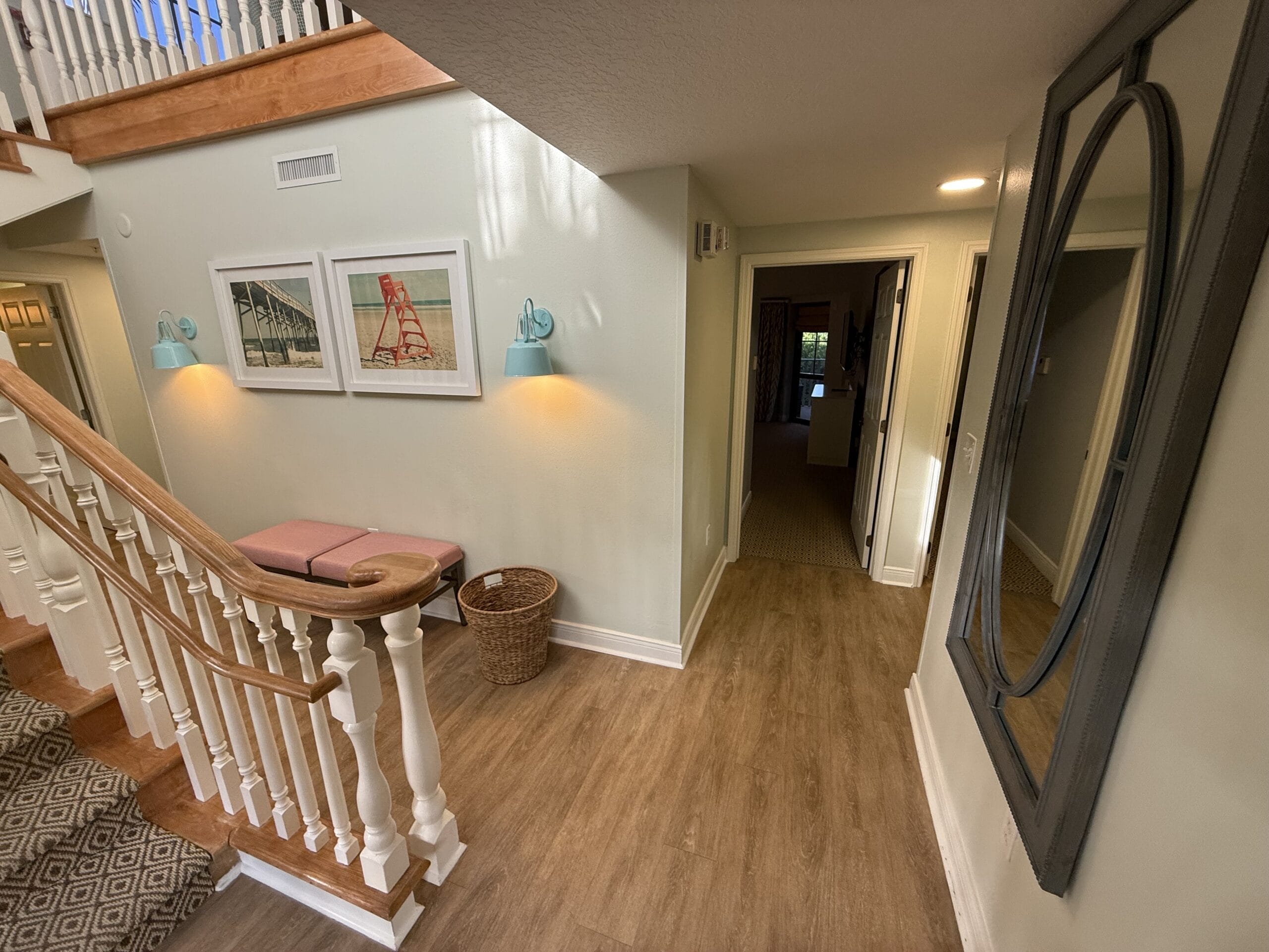 Sunlit hallway in Disney's Vero Beach Resort 3 Bedroom Cottage with wood floors, staircase, Disney wall art, and a round mirror.
