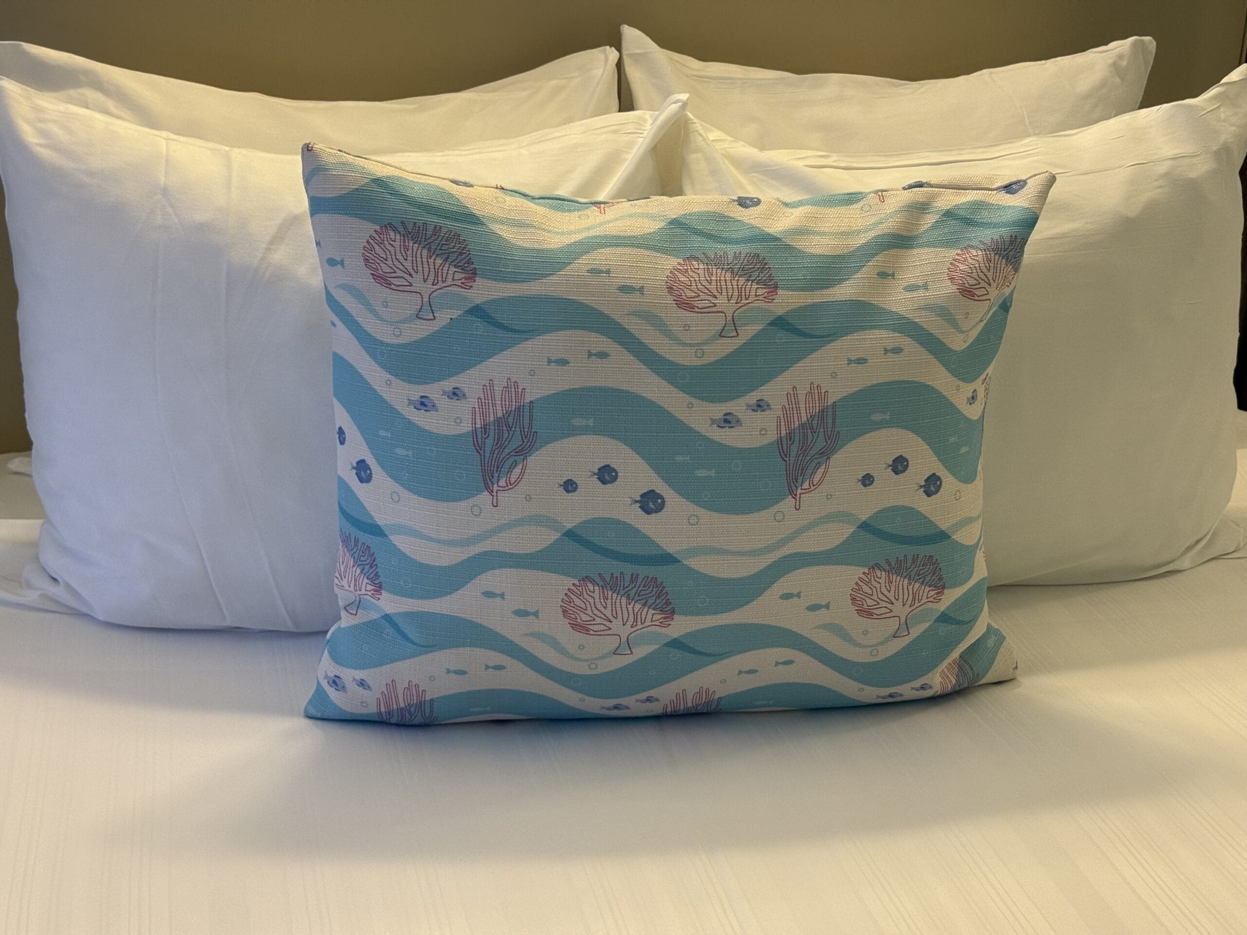 A bed at Disney’s Vero Beach Resort, topped with white pillows and a pillow featuring blue waves, coral, and fish accents in front.