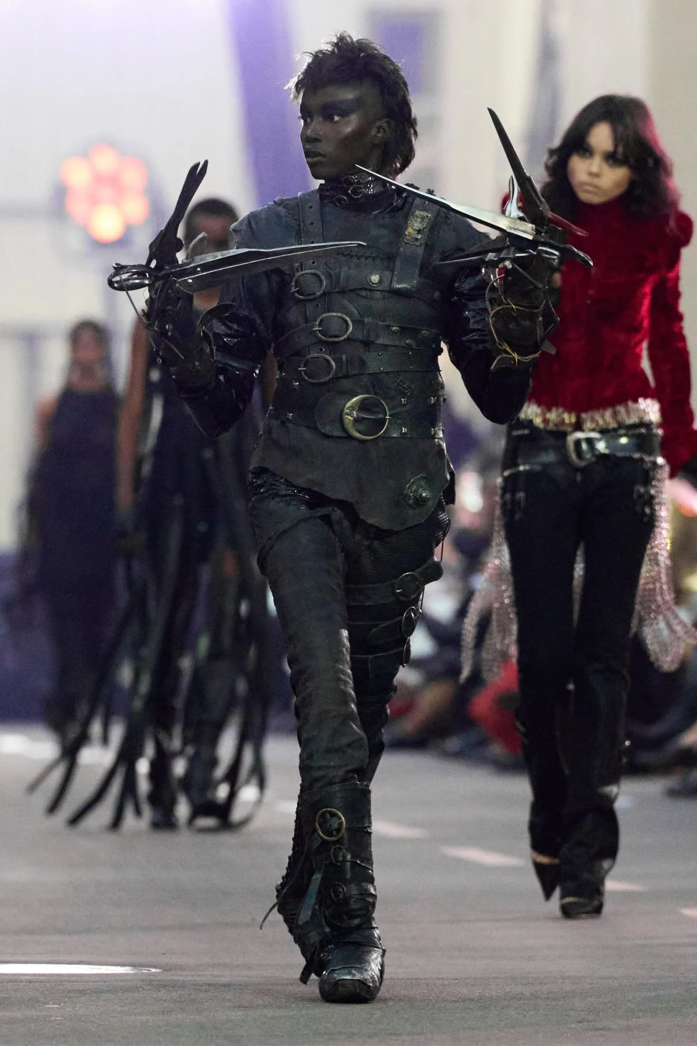 A model in black, Edward Scissorhands-inspired outfit with scissor hands walks the runway, evoking Disney's darker fairytale style.